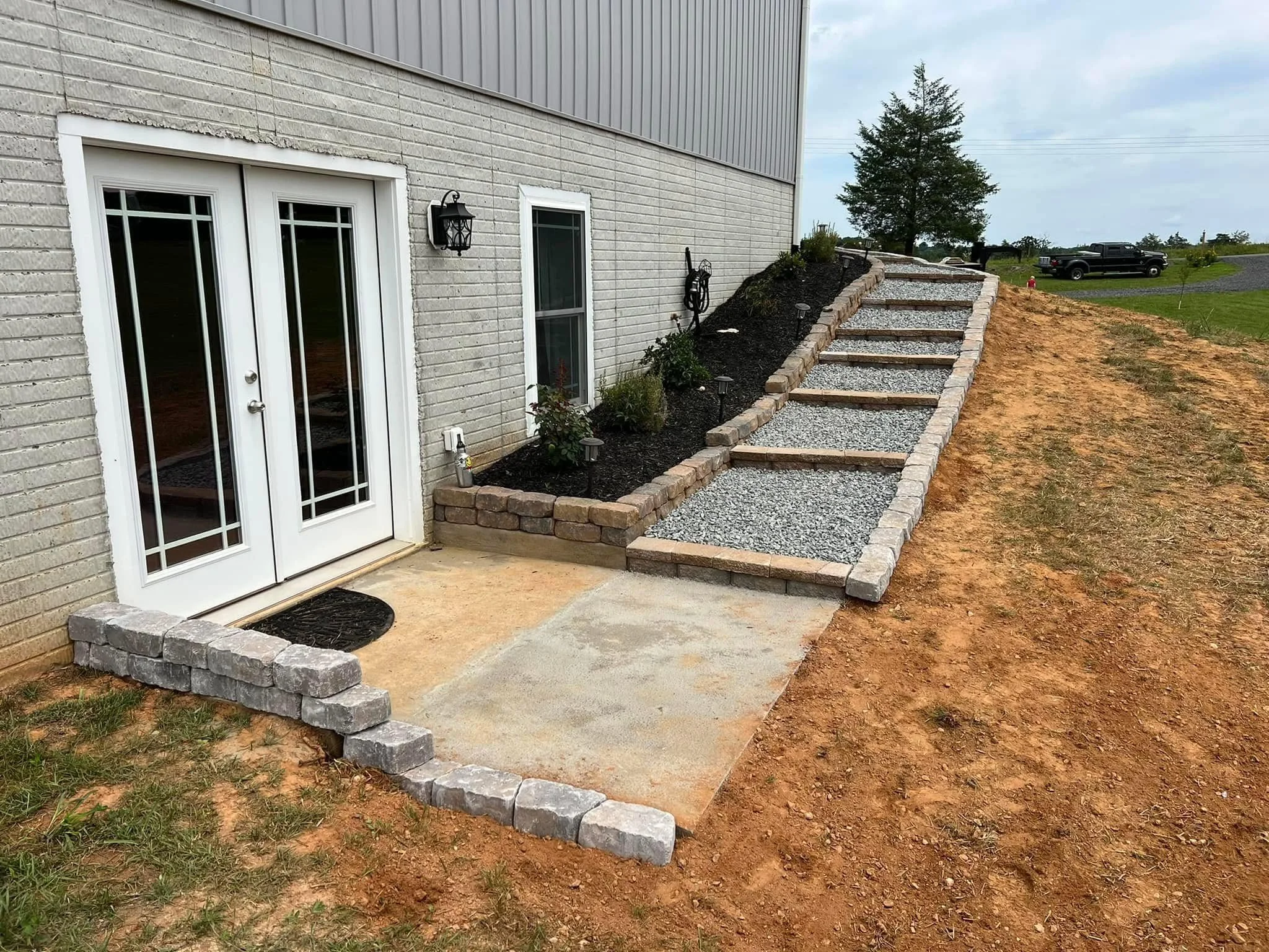 Backyard with white brick house and a landscaped staircase with stone steps and gravel, leading up a small hill, with a pickup truck and open grassy area in the background.