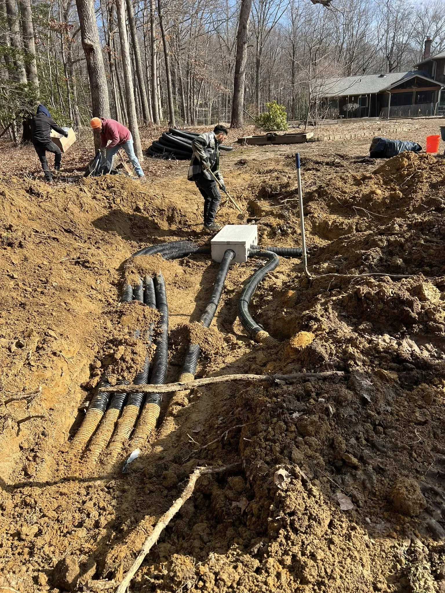Construction workers installing underground header lines for septic system in a dirt excavation site near a wooded area and a house.