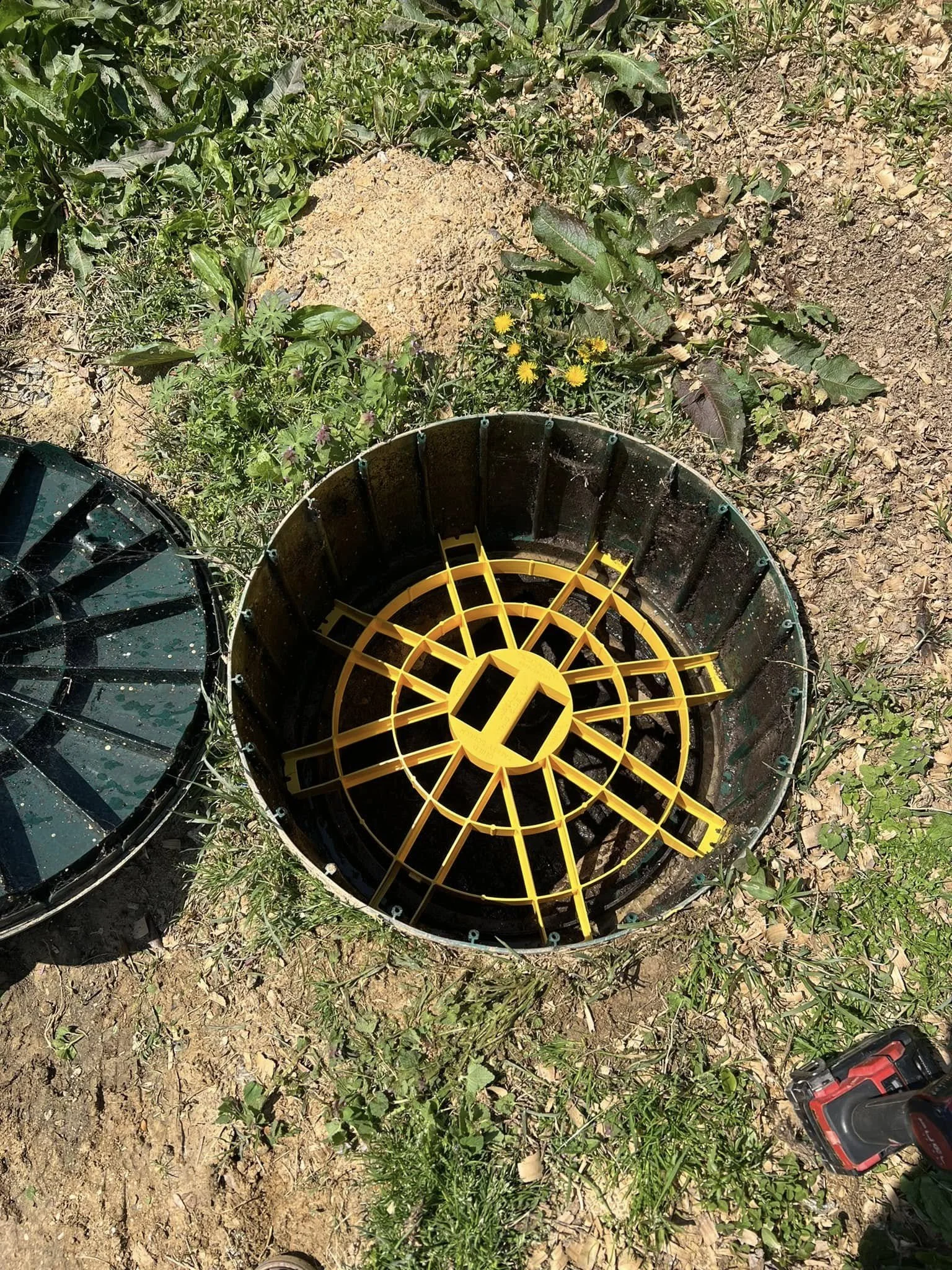 Fall protection added to drainage basin with a yellow grate inside, placed on dry soil with grass and plants around.