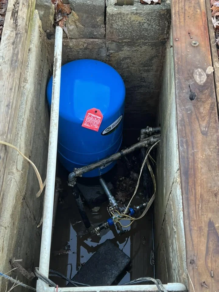 Blue pressure tank installed in a small excavated space with pipes and wires connected.