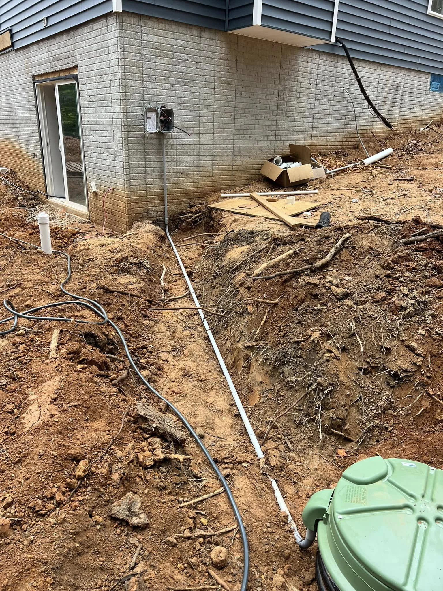 Construction site with exposed dirt, pipes, and electrical wires near the foundation of a building with a brick and siding exterior. An open electrical box and plumbing materials are visible.