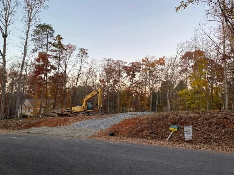 Construction site with a yellow excavator on a dirt and gravel hill, surrounded by trees with autumn foliage, near a residential neighborhood.