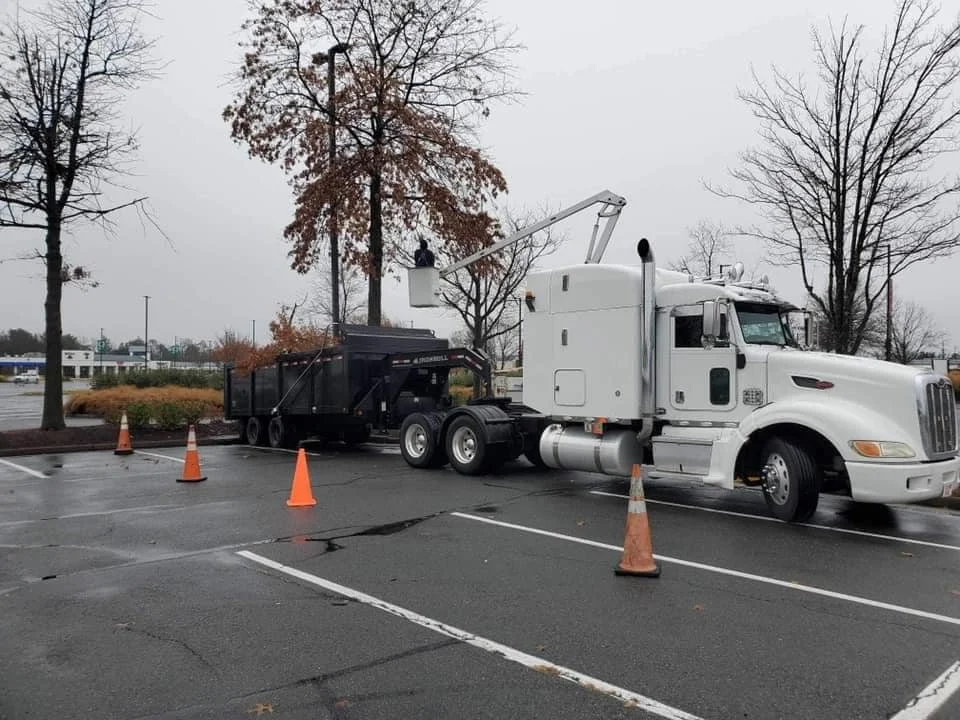 A white semi-truck with a crane attachment is parked in an empty parking lot, nearby orange traffic cones, leafless trees, and an overcast sky.