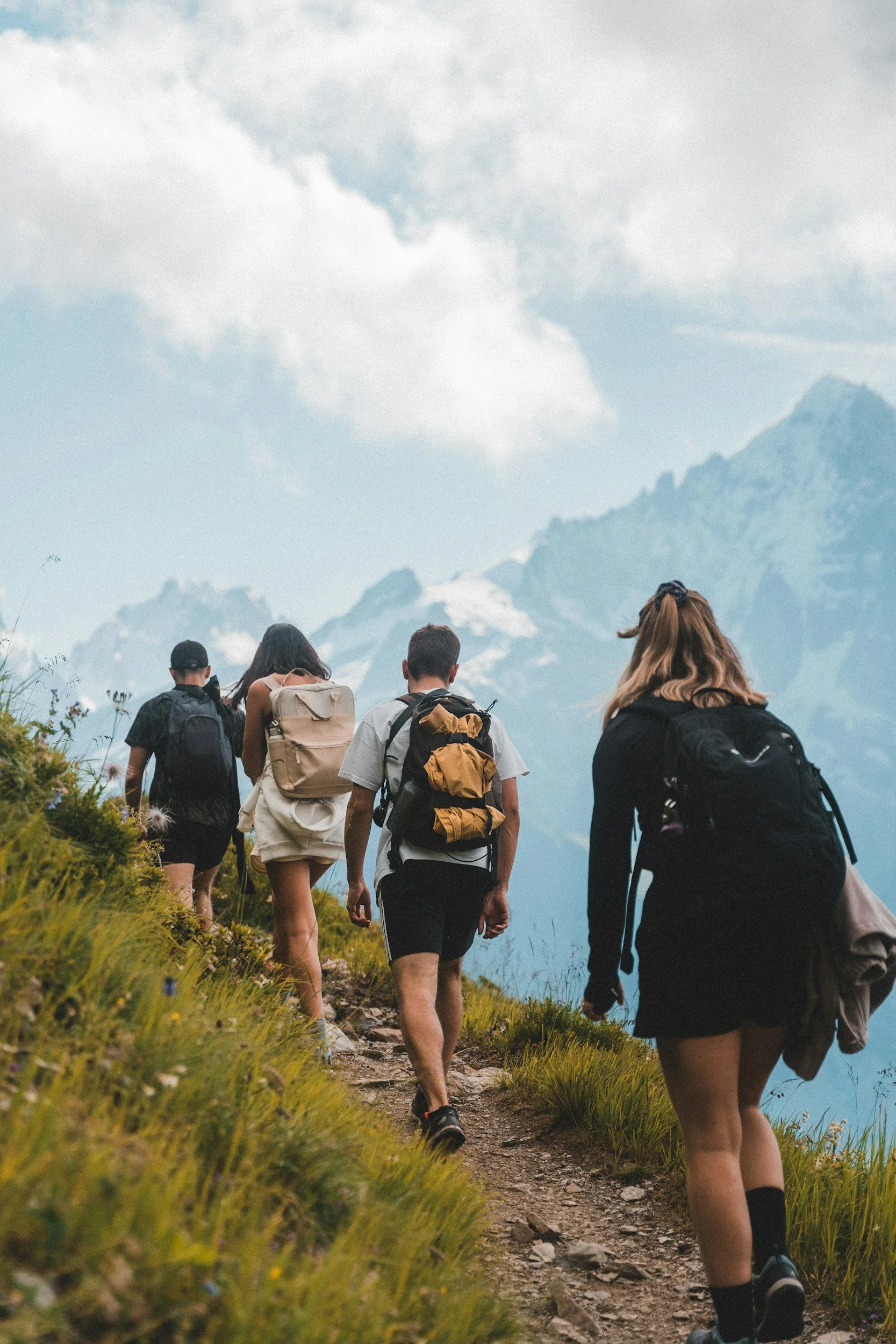 Group of five hikers walking uphill on a mountain trail with snow-capped peaks in the background.