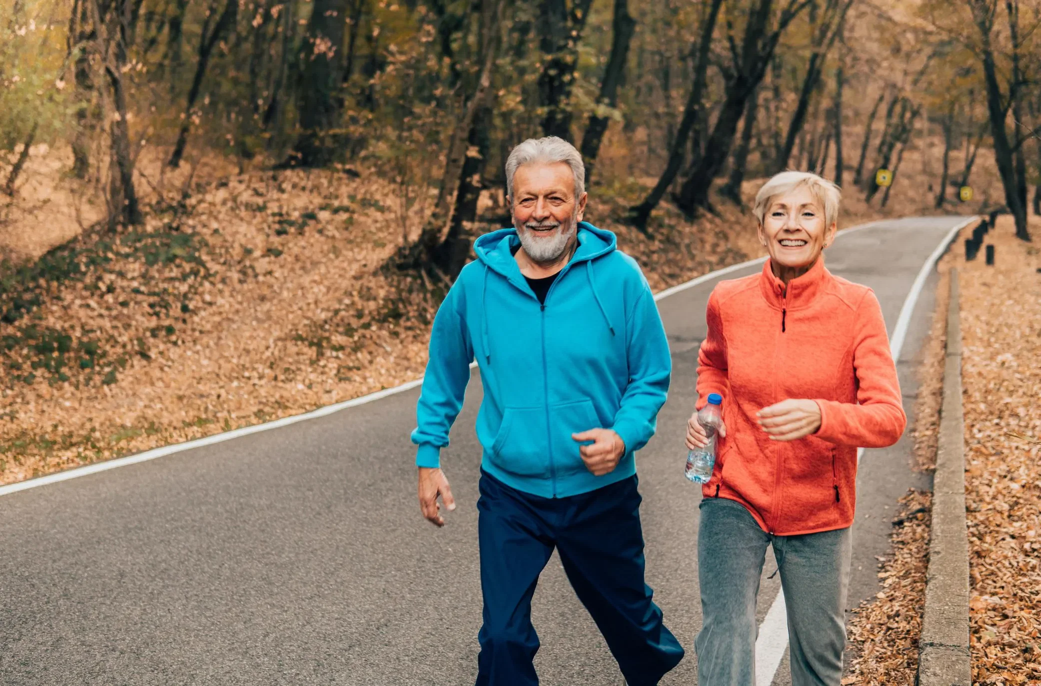 Older man and woman jogging on a paved road surrounded by autumn trees with fallen leaves.