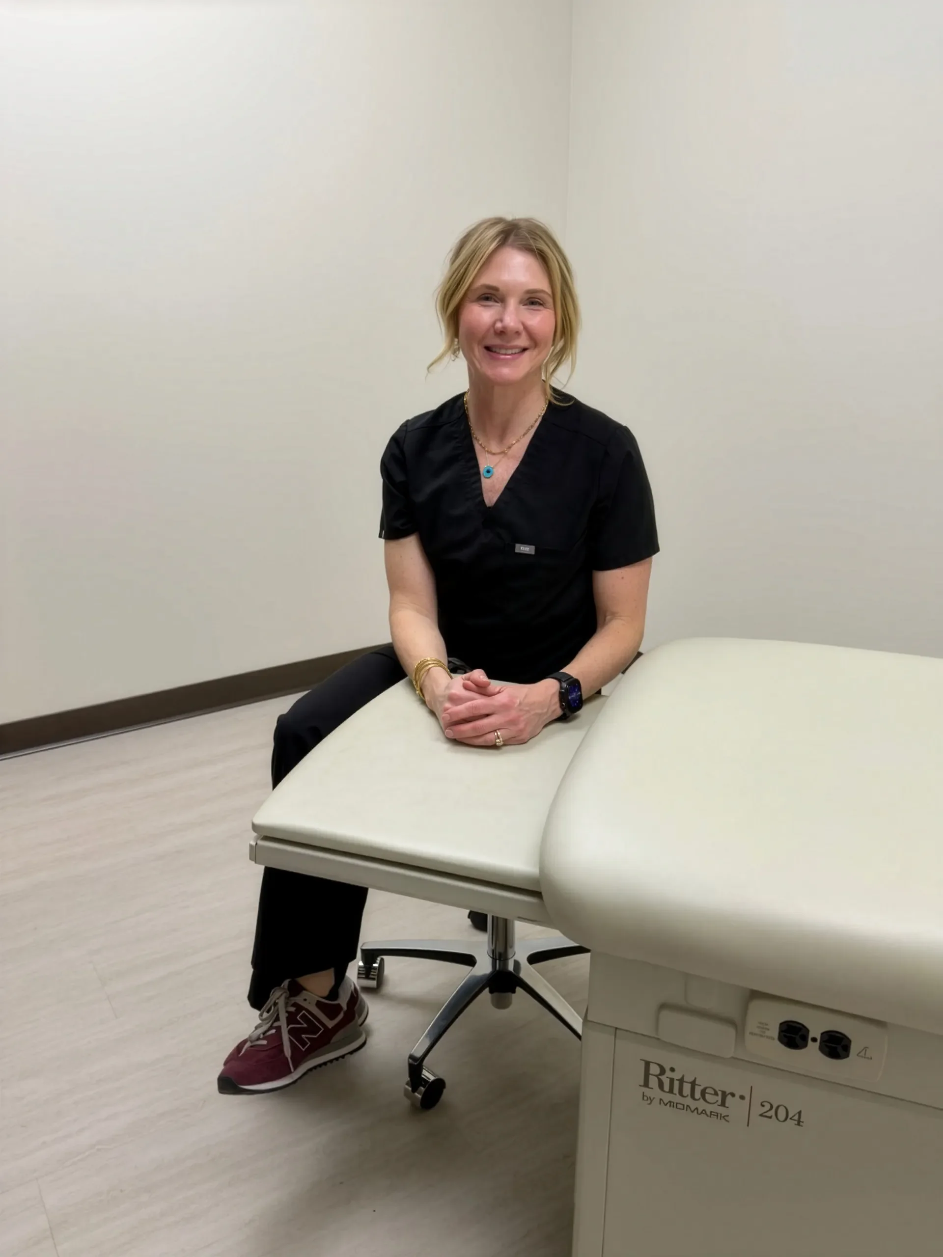 A woman sitting on an examination table in a medical office, wearing black scrubs, sneakers, and jewelry, smiling at the camera.