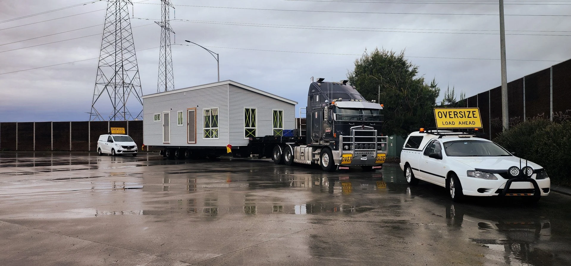 A large flatbed truck carrying a small white building with multiple windows, parked in a lot on a rainy day.