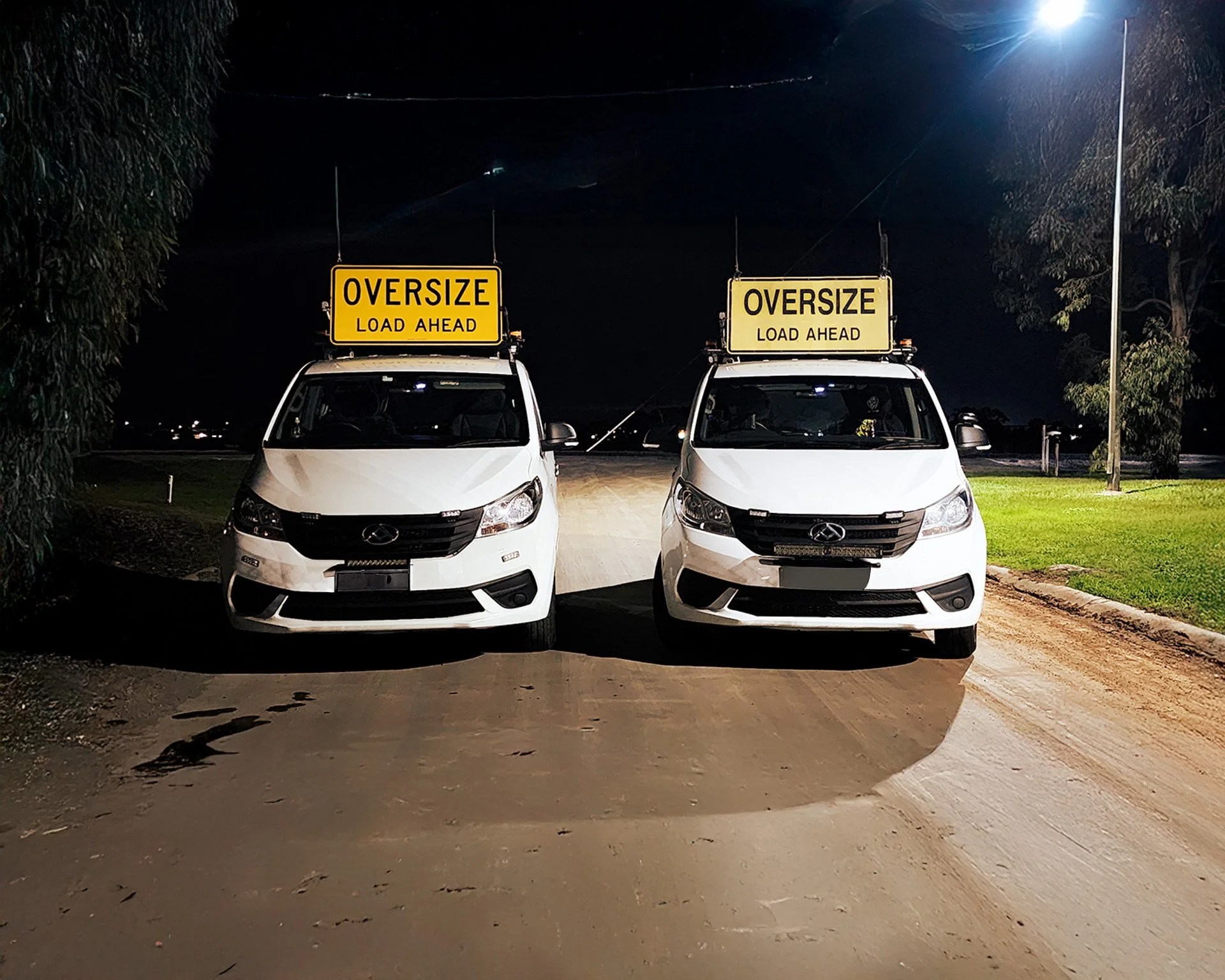 Two white NC3 OSOM vans parked at night under overhead signs that read "OVERSIZE LOAD AHEAD." The scene is lit by a streetlamp on the right side, with trees and a grassy area visible in the background.