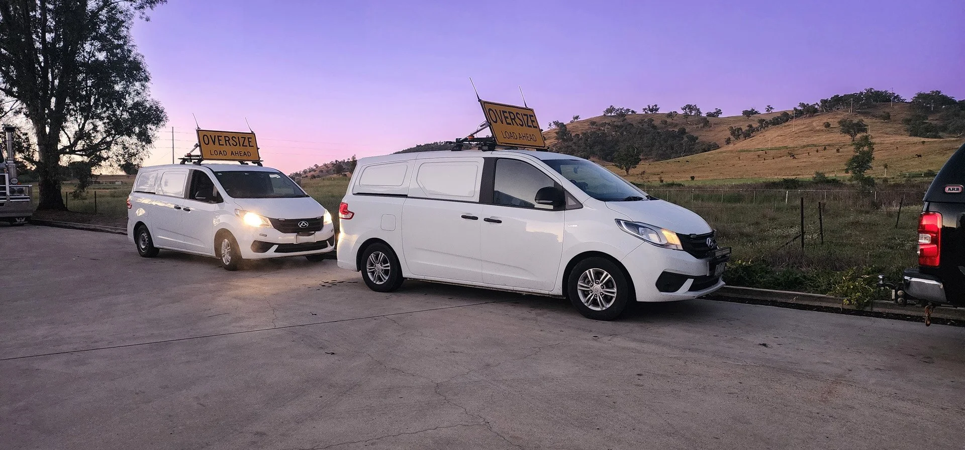Two white NC3 OSOM vans with oversized load signs parked on a concrete surface, with a landscape of hills and trees in the background during dusk.
