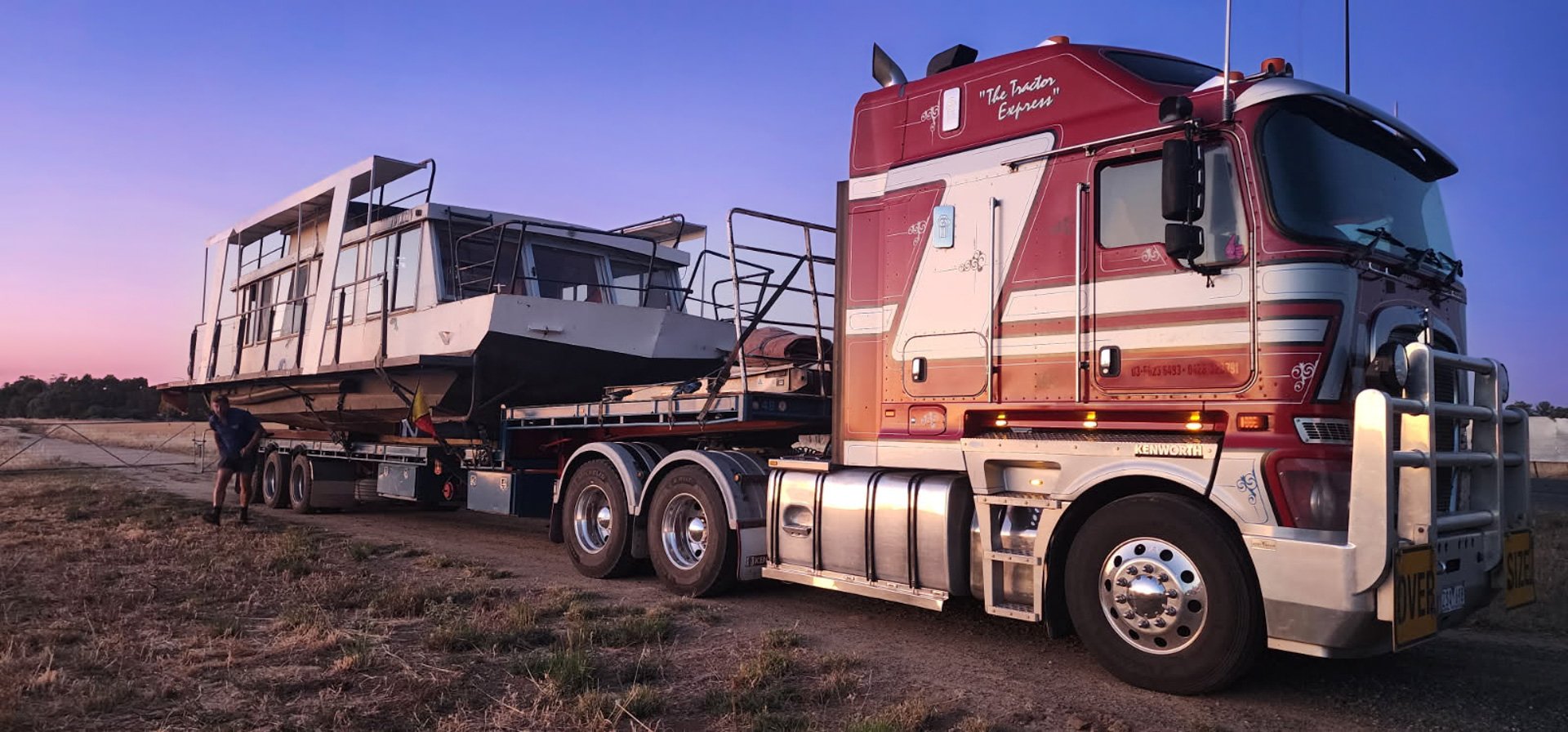 A large red semi-truck with a white stripe, hauling a boat on a trailer in an open field at sunset.