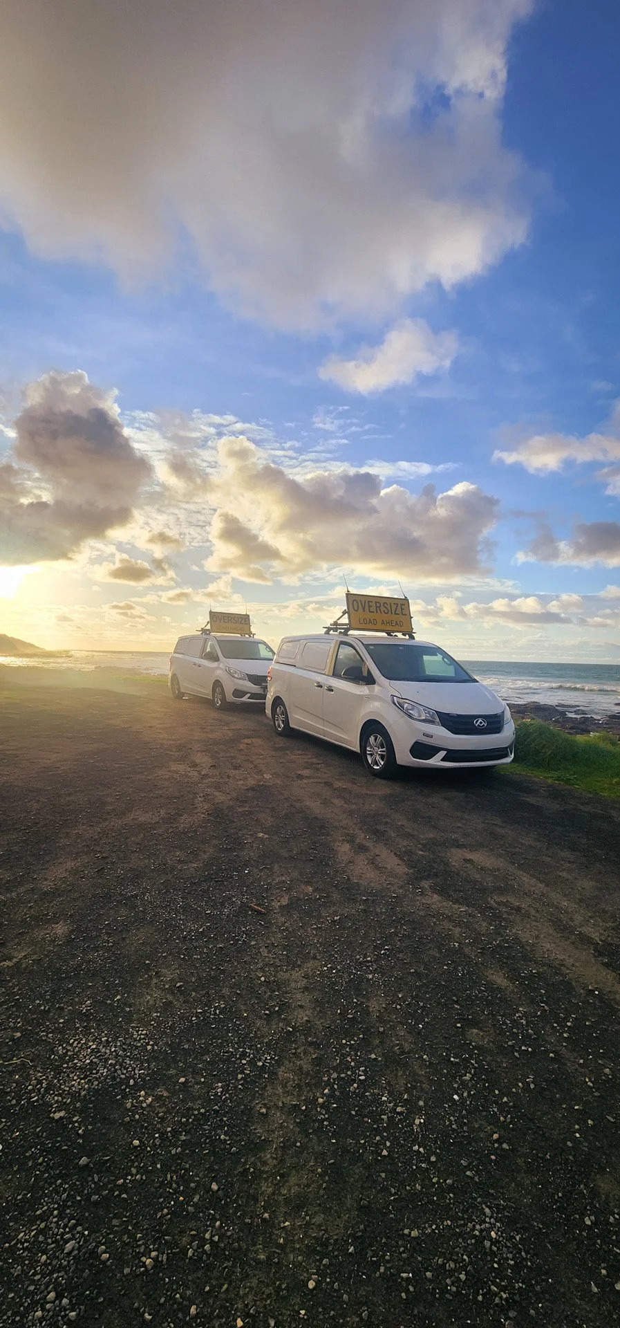 Two white NC3 OSOM vans parked on a dirt path near the coast, both with oversize load signs on their roofs, under a cloudy sky at sunset.