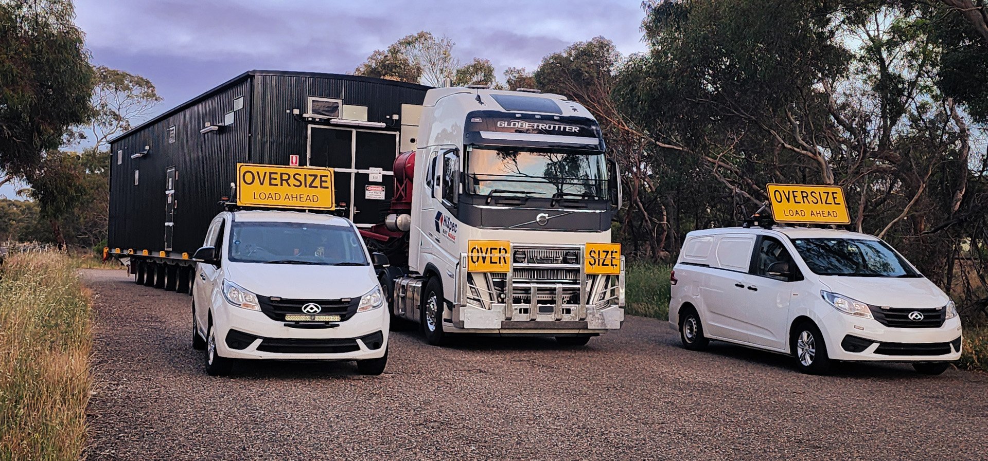 Two white NC3 OSOM vehicles with 'OVERSIZE LOAD AHEAD' signs parked beside a large truck with a sign that says 'OVER SIZE'. The scene is outdoors on a gravel road with trees and a cloudy sky in the background.