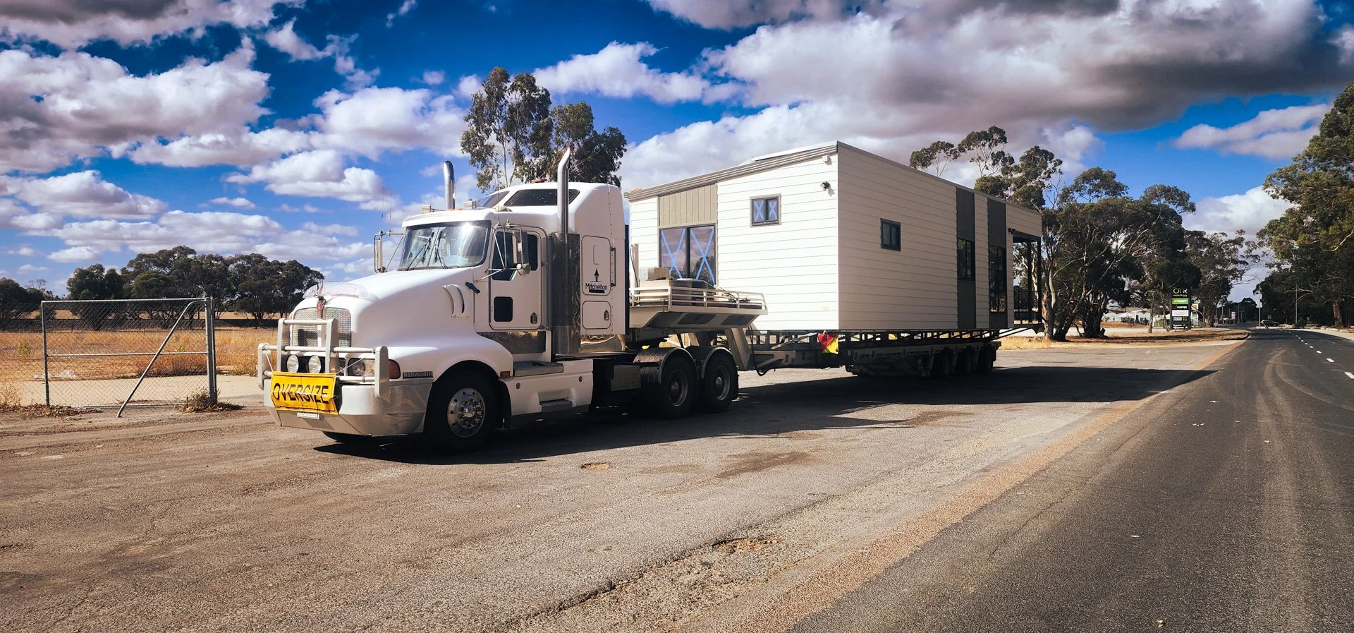 A large white semi-truck with a house on its flatbed trailer driving on a rural road under a partly cloudy sky.