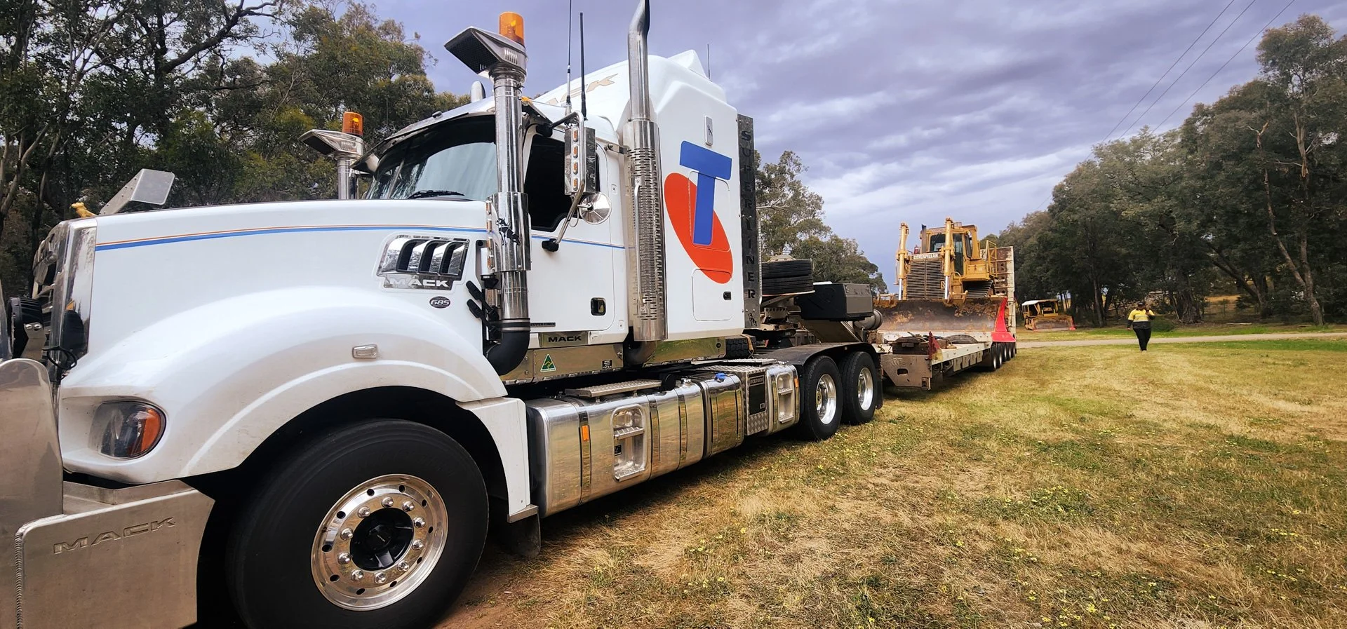 A large white Mack truck hauling construction equipment, including a bulldozer, on a trailer. The scene takes place outdoors in a grassy area with trees, and two people are walking in the background.