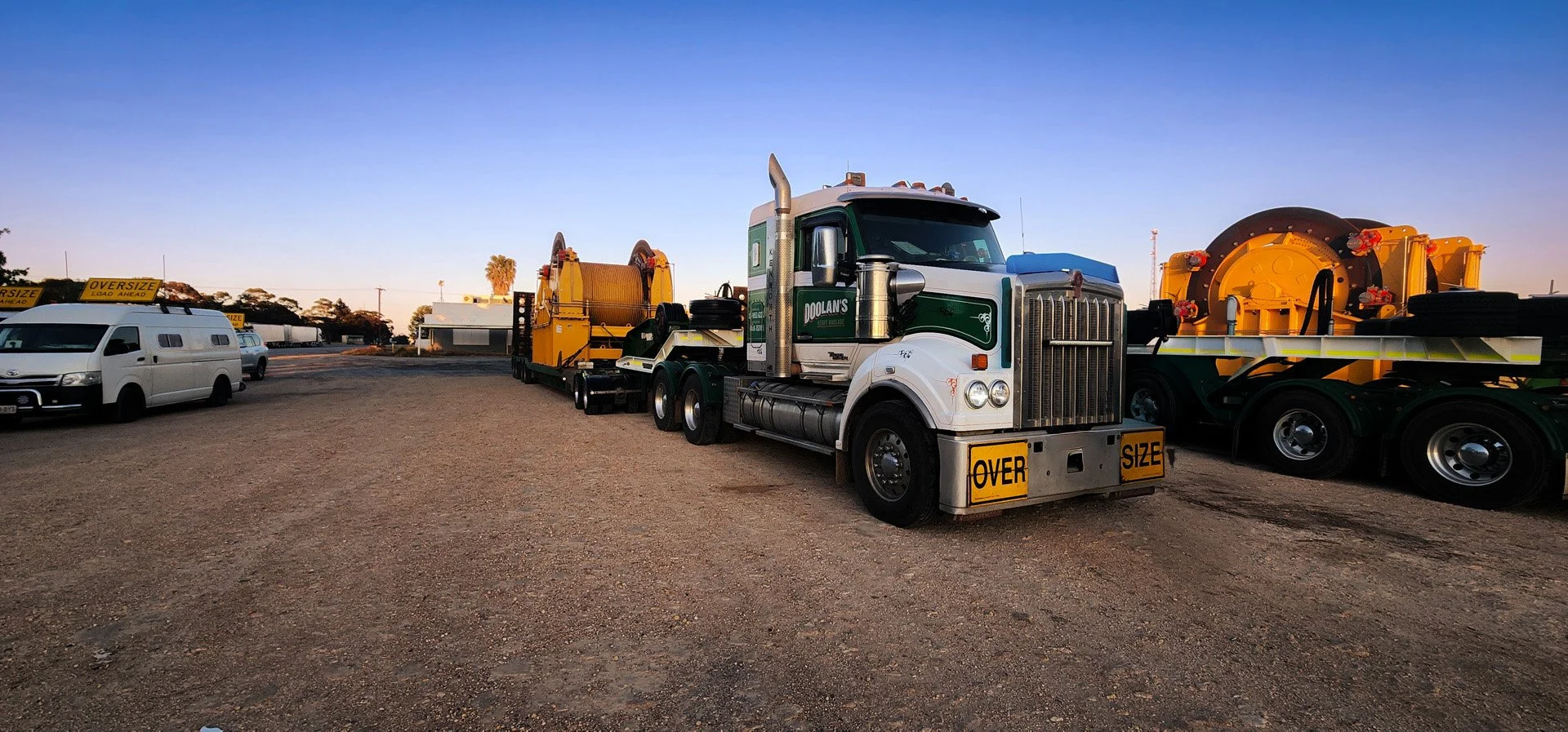 A large semi-truck parked in a lot with oversized load signs, connected to a flatbed trailer carrying industrial equipment, with white vans and other vehicles nearby, under a clear blue sky.