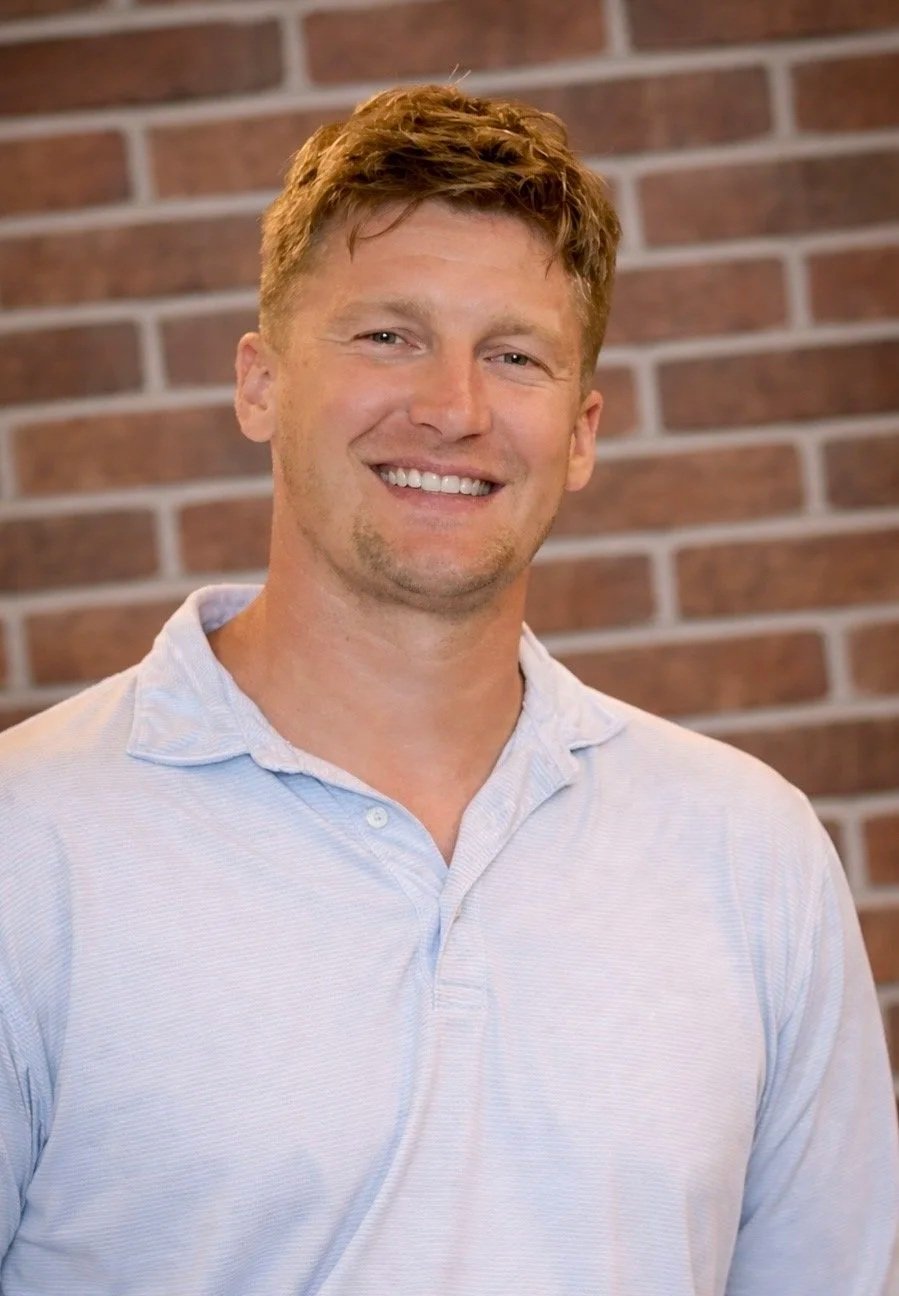 A smiling man with short, curly reddish hair wearing a light gray polo shirt, standing in front of a brick wall.