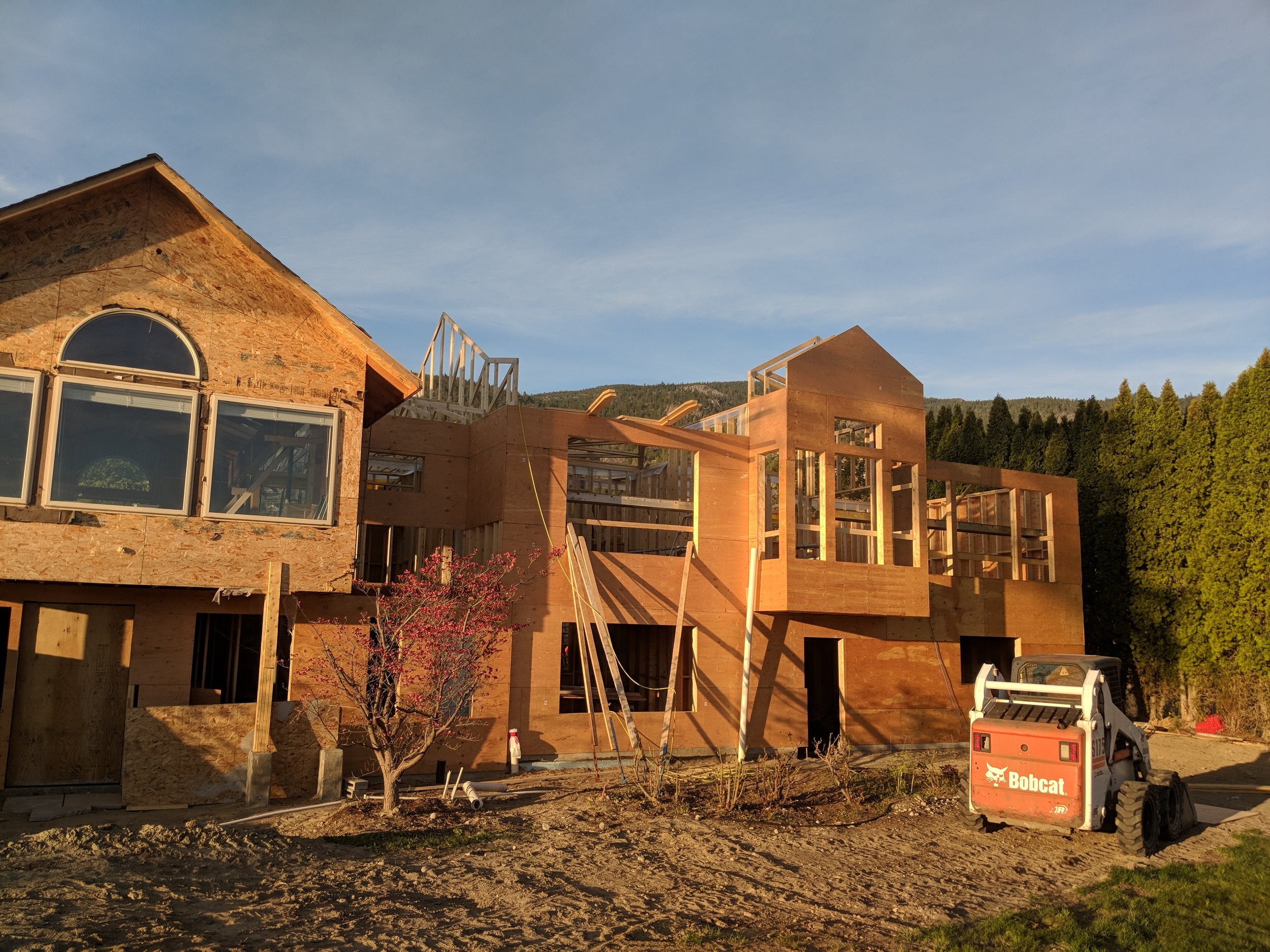 A house under construction with wooden framing and brick exterior, a small tree in front, and a Bobcat compact loader on the right side.