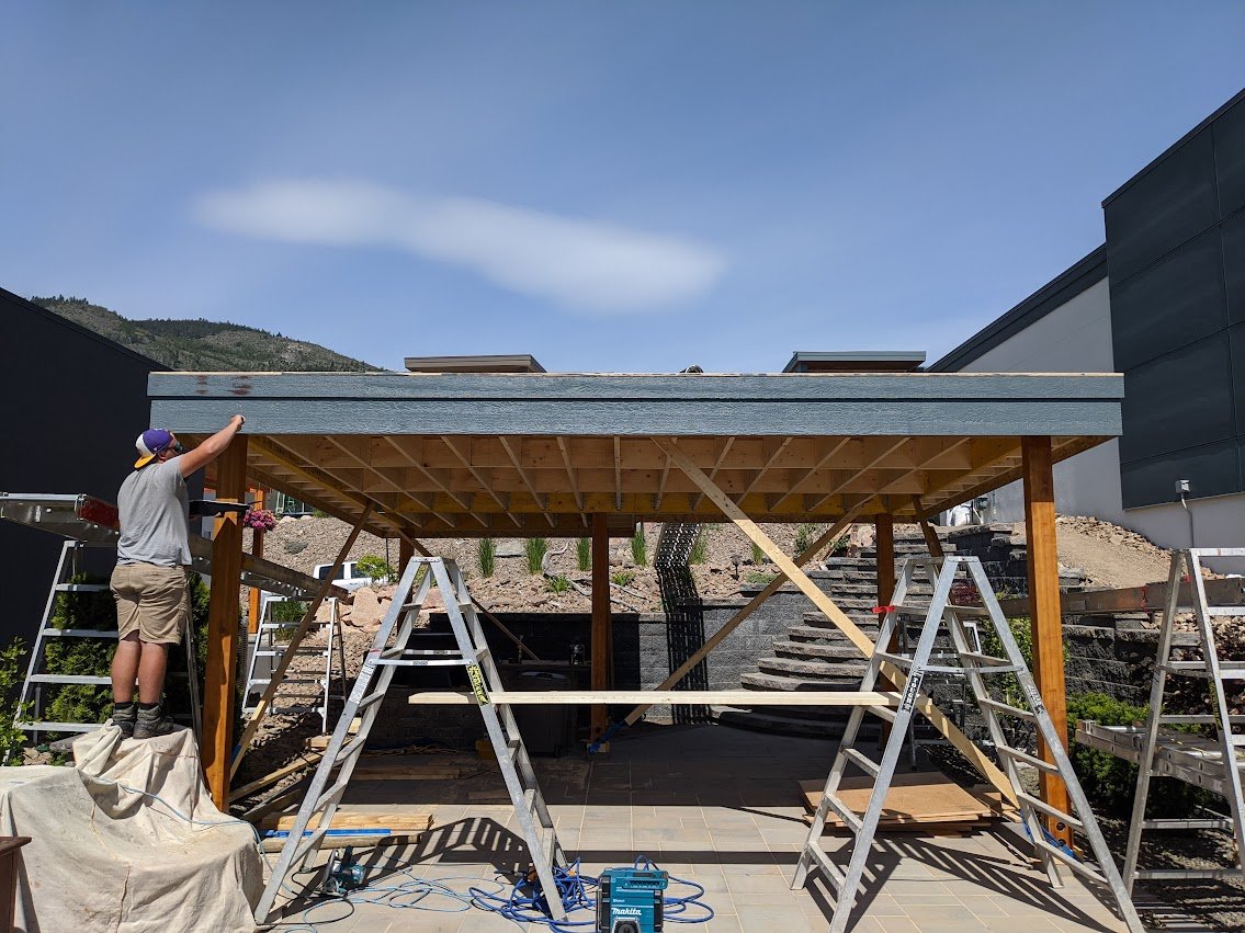 A construction worker working on a wooden structure outdoors, with ladders, tools, and a partly built roof in front of a backdrop of mountains and modern buildings.