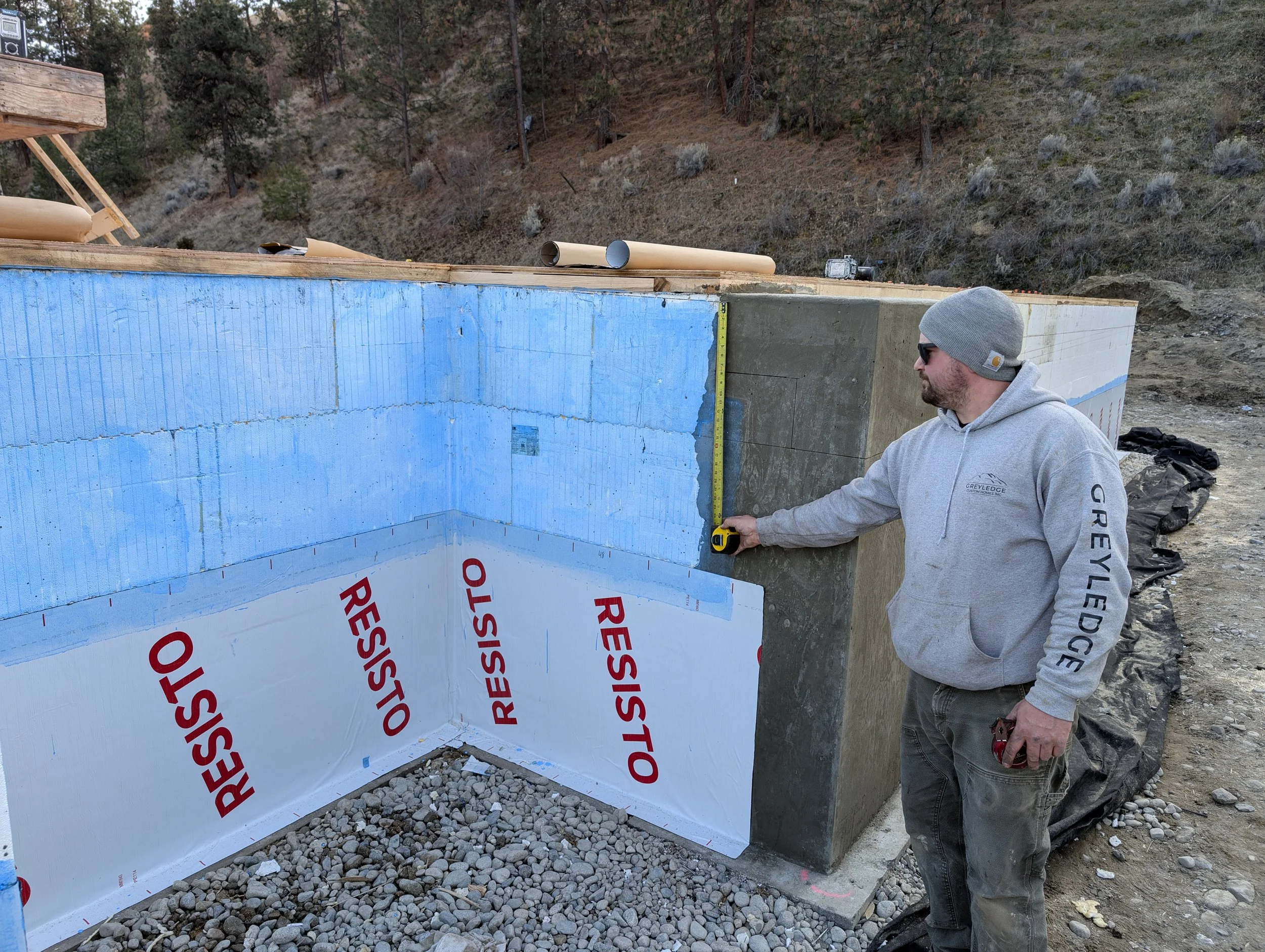 A man wearing sunglasses, a gray beanie, a gray hoodie, and gray pants is measuring a wall at a construction site with a tape measure. The wall is blue insulation with red lettering that reads 'RESISTO'. The site is outdoors with a rocky ground and a hillside with dry grass and scattered trees in the background.