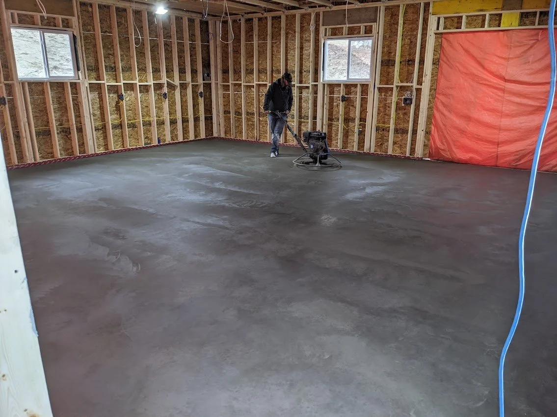 A person working on a concrete slab in an unfinished room of a building under construction, with exposed wooden framing, small windows, and construction materials around.