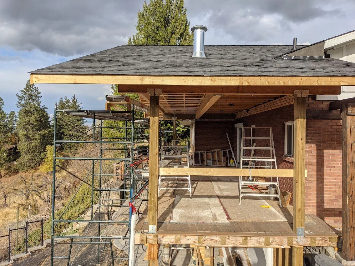 Construction of a wooden deck extension on the back of a brick house. Scaffolding surrounds the area, and there are ladders and construction tools present.