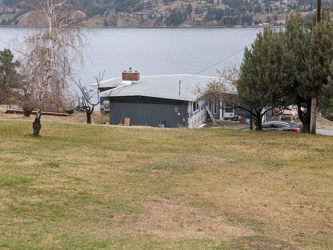 A lakeside house with gray siding and a white roof, surrounded by trees with a lake in the background.