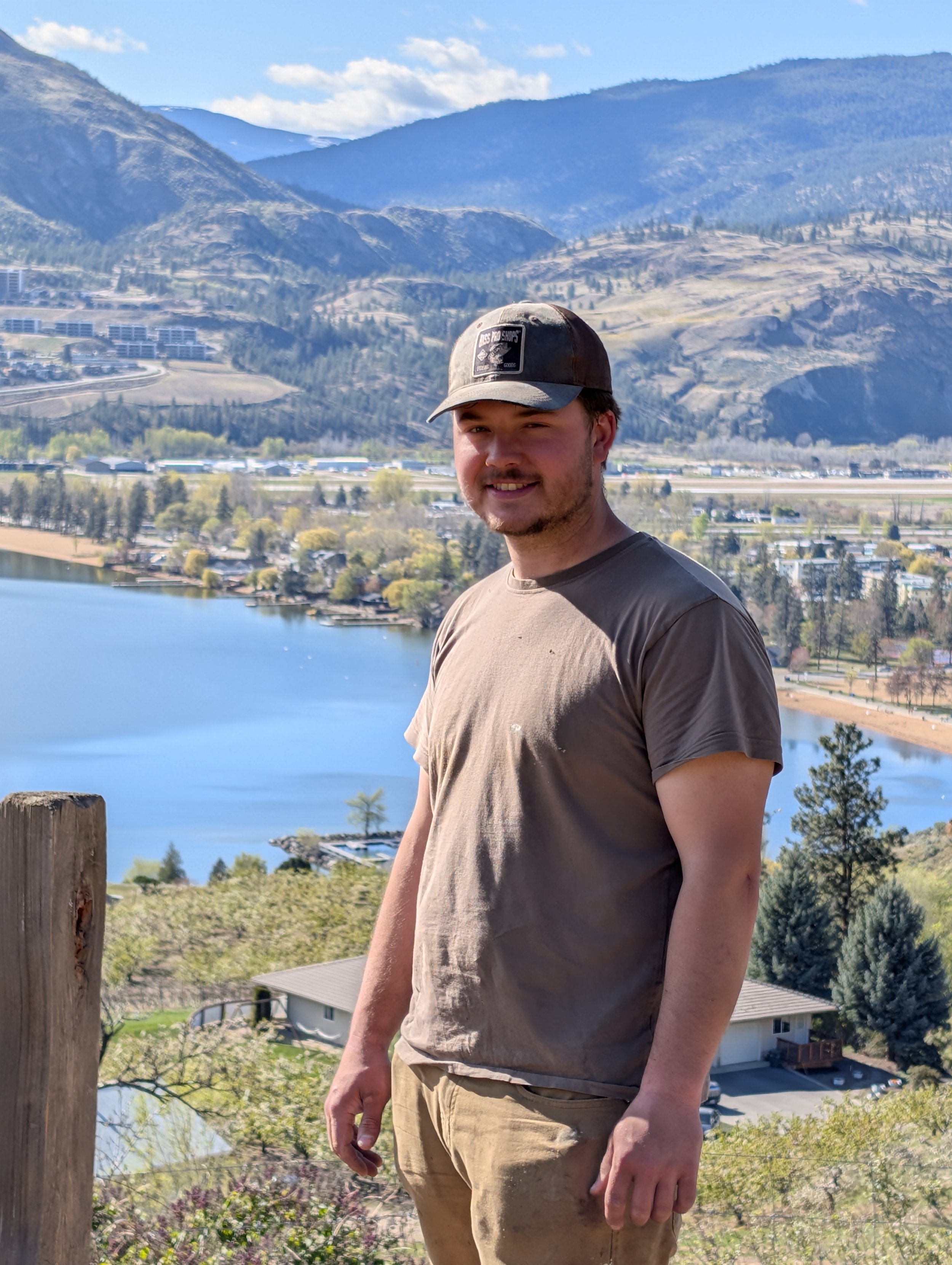 A young man smiles at the camera while standing outdoors with mountains, a lake, trees, and houses in the background on a sunny day.