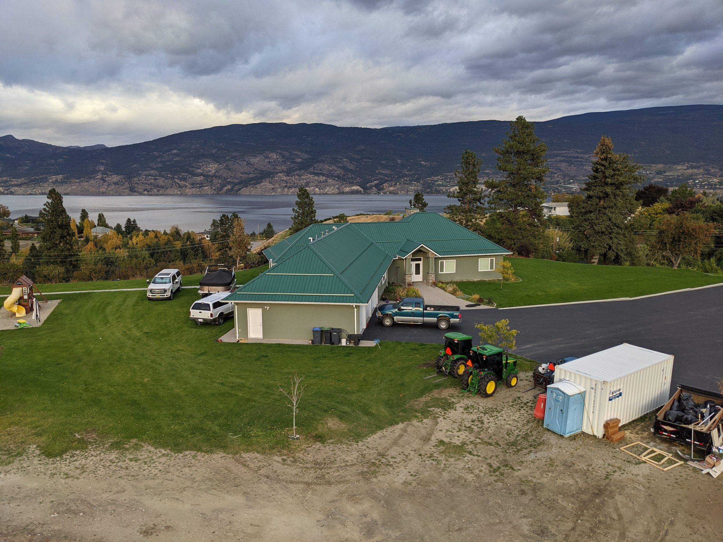 A scenic view of a lake surrounded by mountains under a cloudy sky. In the foreground, there are residential houses with green roofs, a few parked cars, and a backyard with a small playground, a tractor, and shipping containers.