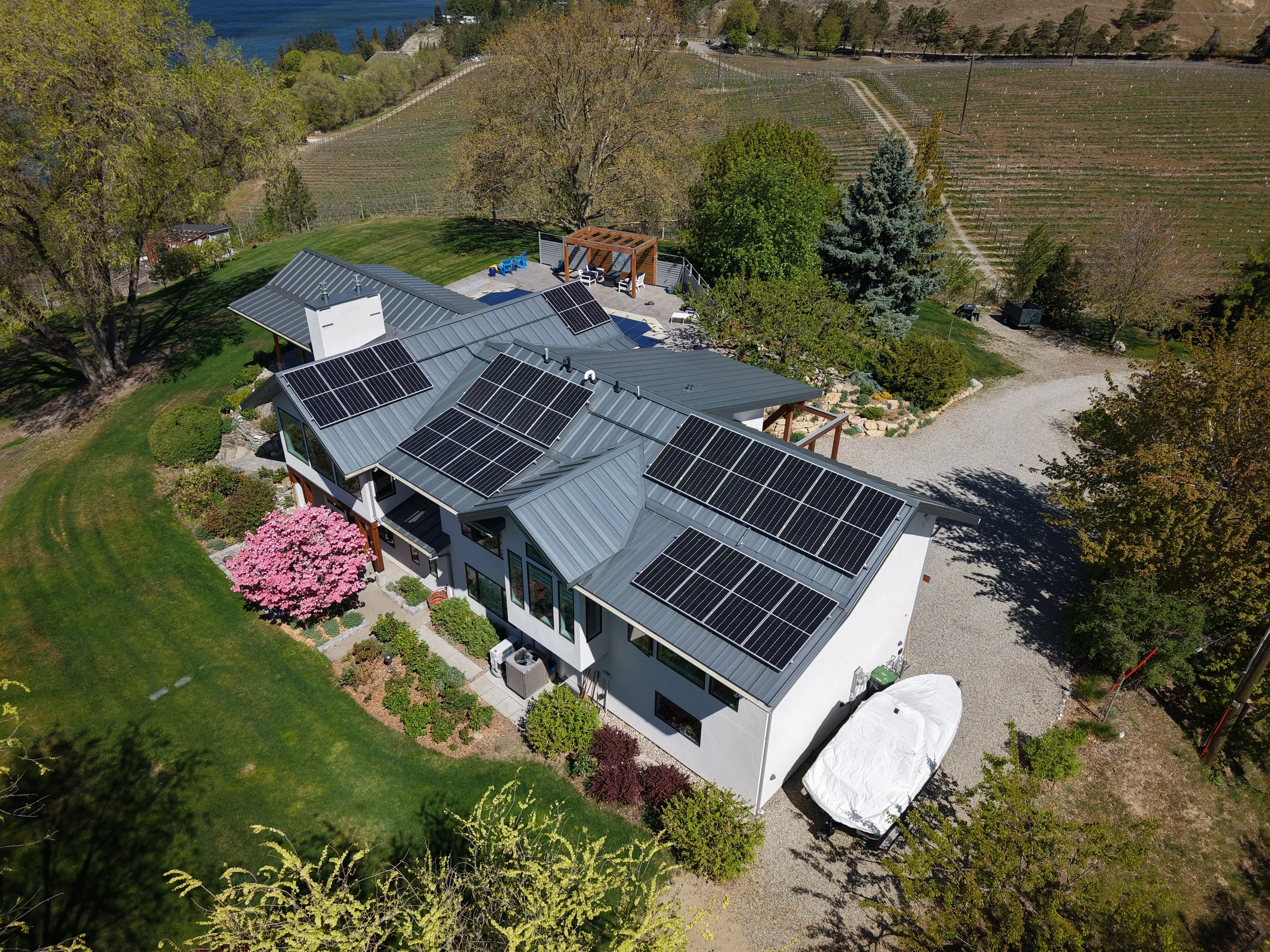An aerial view of a house with solar panels on its roof, set in a lush yard with trees and outdoor seating, surrounded by fields and vineyards.