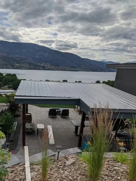 A backyard patio with outdoor furniture near a lake and mountain backdrop under a cloudy sky.