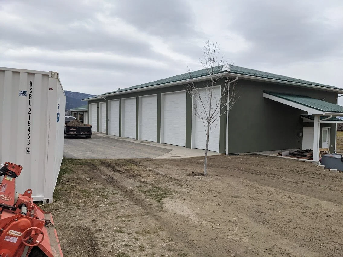 A large building with a green metal roof and multiple white garage doors, a small bare tree in front, and a cloudy sky overhead. There is a small driveway and exposed dirt around the building, with construction or storage equipment nearby.