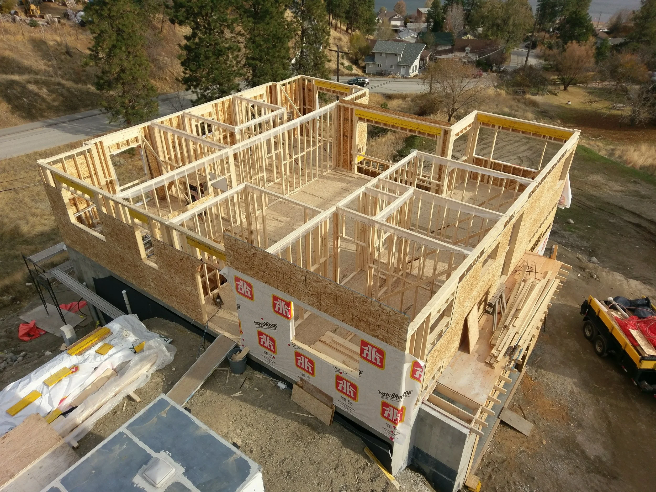 A multi-story house under construction with exposed wooden framing, insulation panels, and construction materials on the ground, in a suburban area with trees, houses, and a road in the background.
