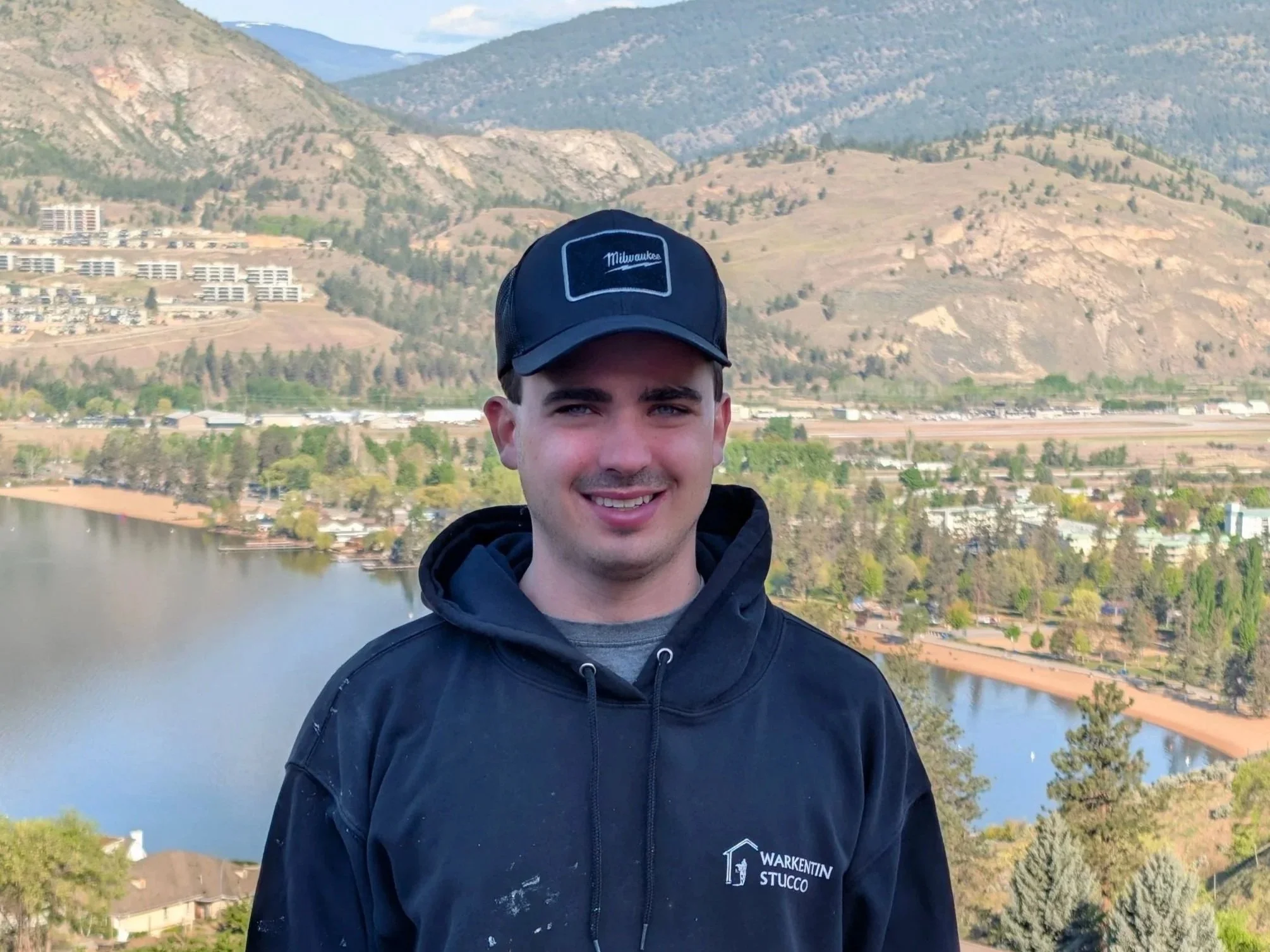 A young man wearing a dark hoodie and a cap with 'Milwaukee' on it, smiling outdoors with a river, houses, and mountain landscape in the background.