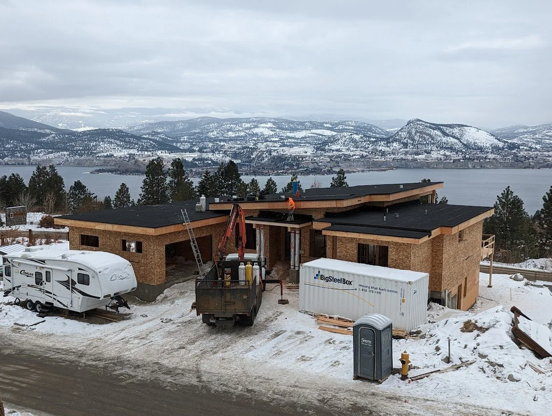 Under construction house with partially completed roof, construction equipment, a trailer, and a portable toilet in a snowy landscape near a lake and mountains.