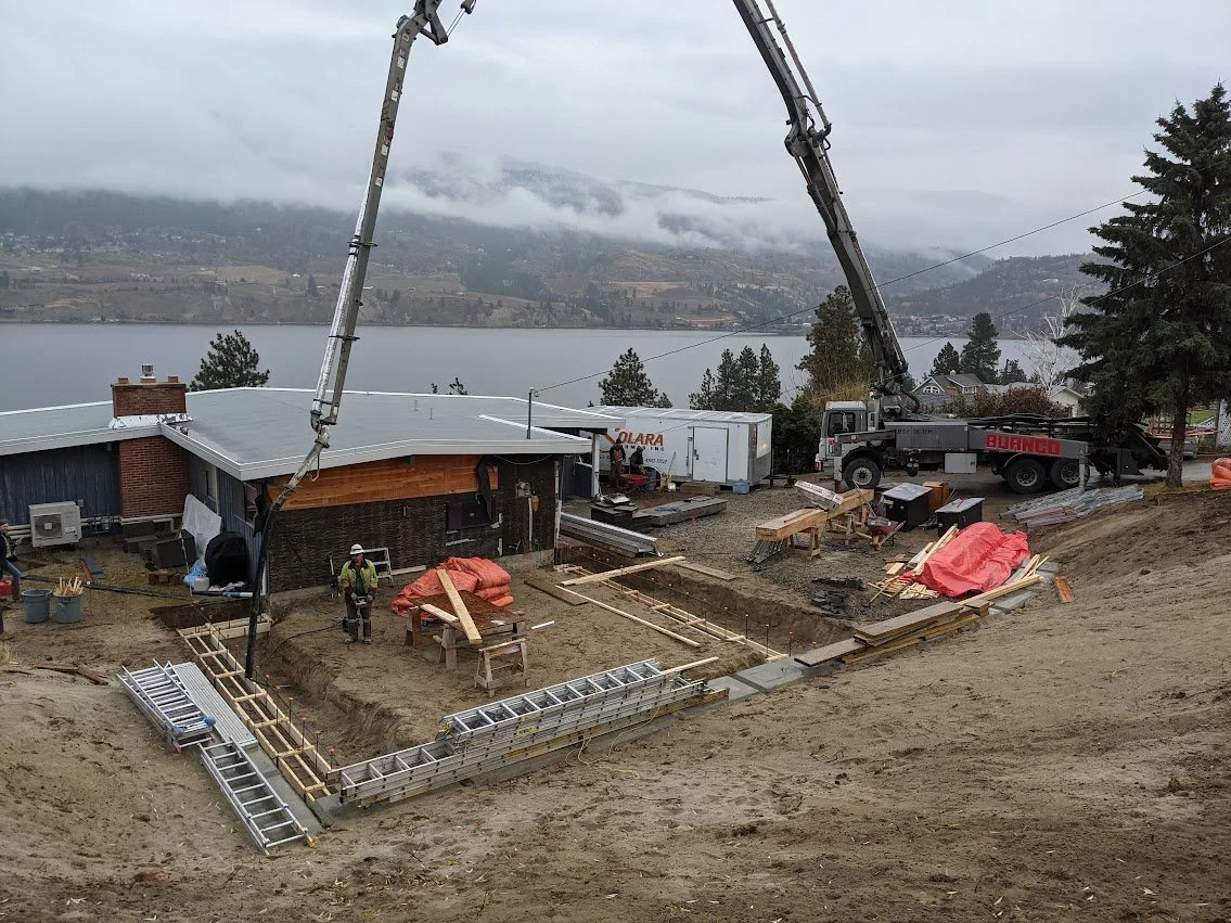 Construction site on a sloped lot with a lake and mountains in the background, showing a concrete pump truck, workers, and building materials for a house.