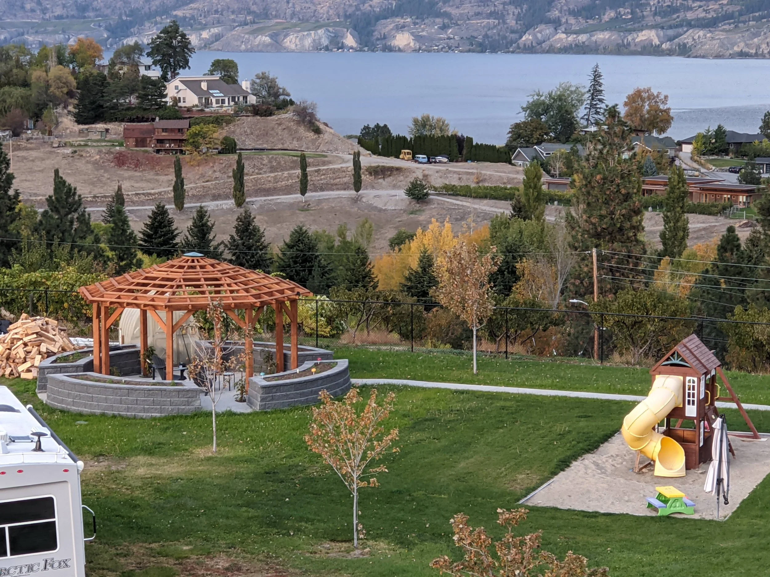 A backyard with a yellow slide, a small playhouse, a wood-fired oven, and a partially constructed wooden gazebo, with a view of trees, hills, and a lake in the background.