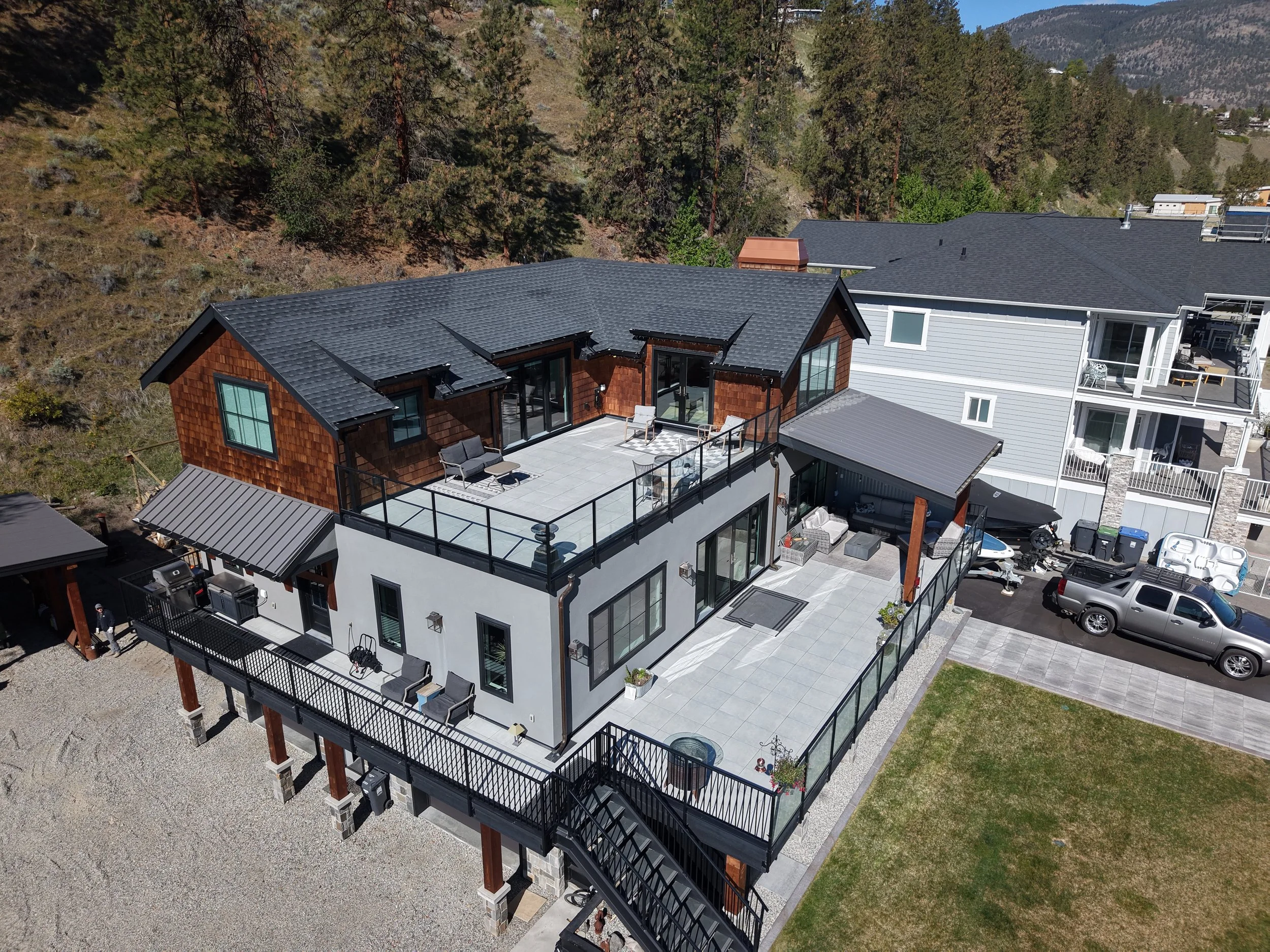 Aerial view of modern residential buildings with balconies, parking area, and a backyard patio surrounded by trees and mountains in the background.