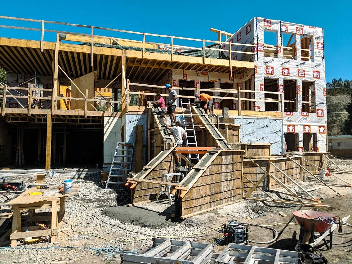Construction workers building a multi-story house with wood framing, scaffolding, and a concrete staircase outdoors under a blue sky.