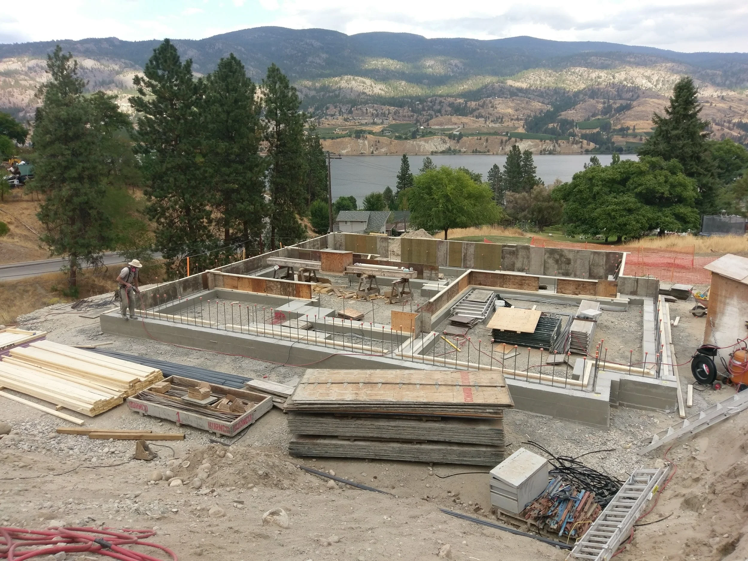 Construction site with concrete foundation being built, surrounded by construction materials, trees, hills, and a lake in the background.