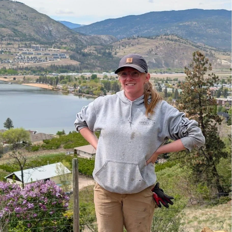 A woman smiling outdoors with hands on hips, wearing a gray hoodie, black cap, and glasses, standing in a scenic landscape with mountains, a lake, trees, and houses in the background.