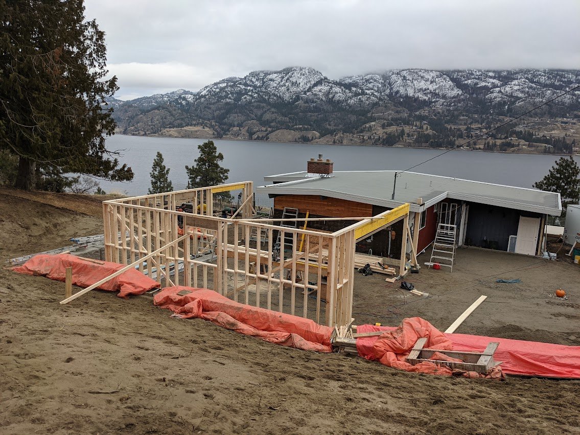 Construction site in progress with a partially built wooden frame on a slope, overlooking a lake and mountains in the background.