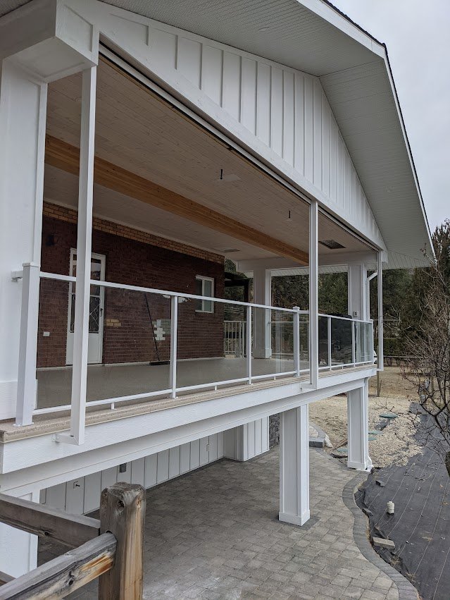 A house with a large, open porch featuring a glass railing, brick wall, and wooden ceiling, with a paved walkway underneath.
