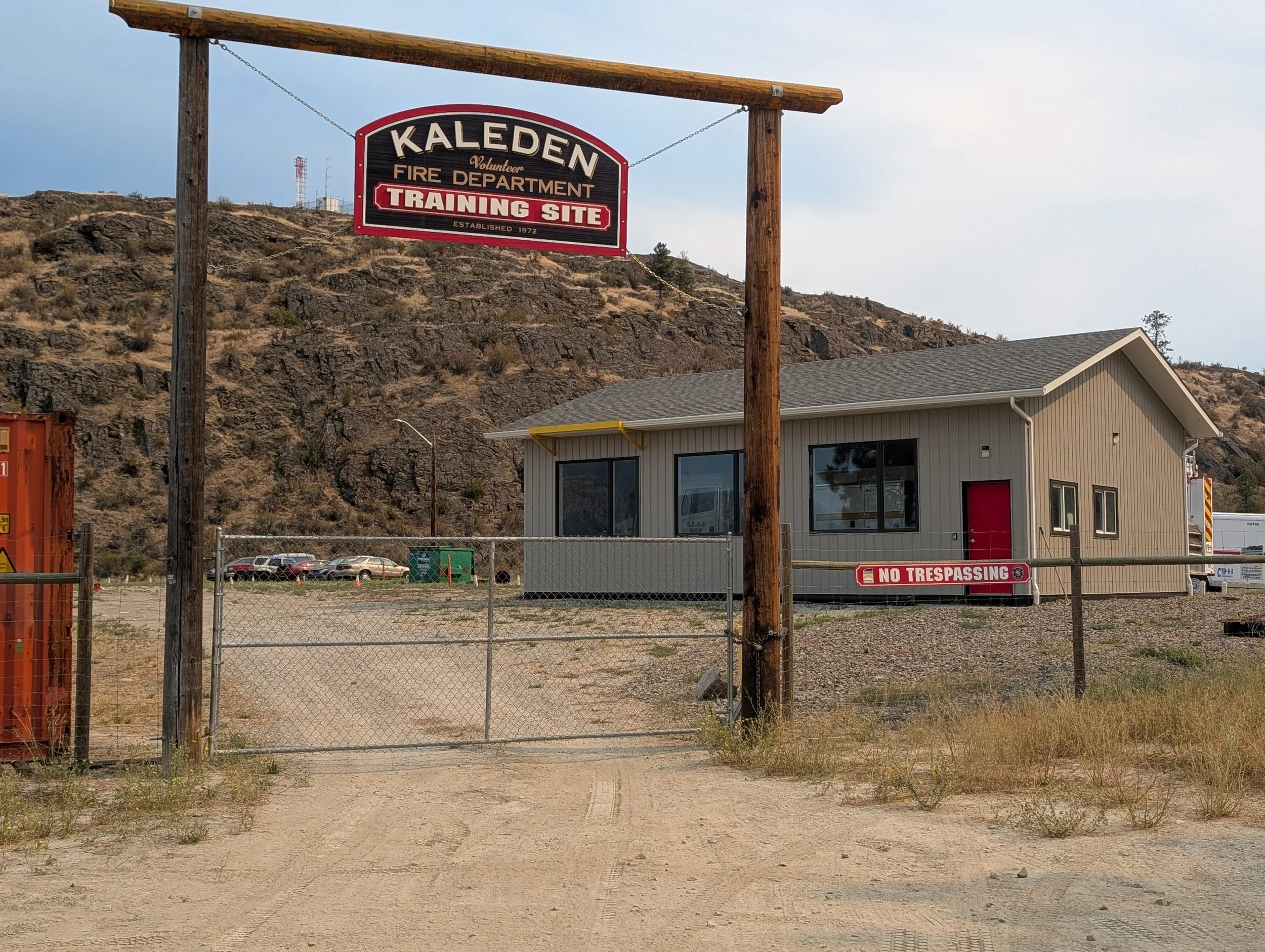 Sign for Kaleden Fire Department training site established in 1972, with a building and a landscape of hills and rocks in the background.