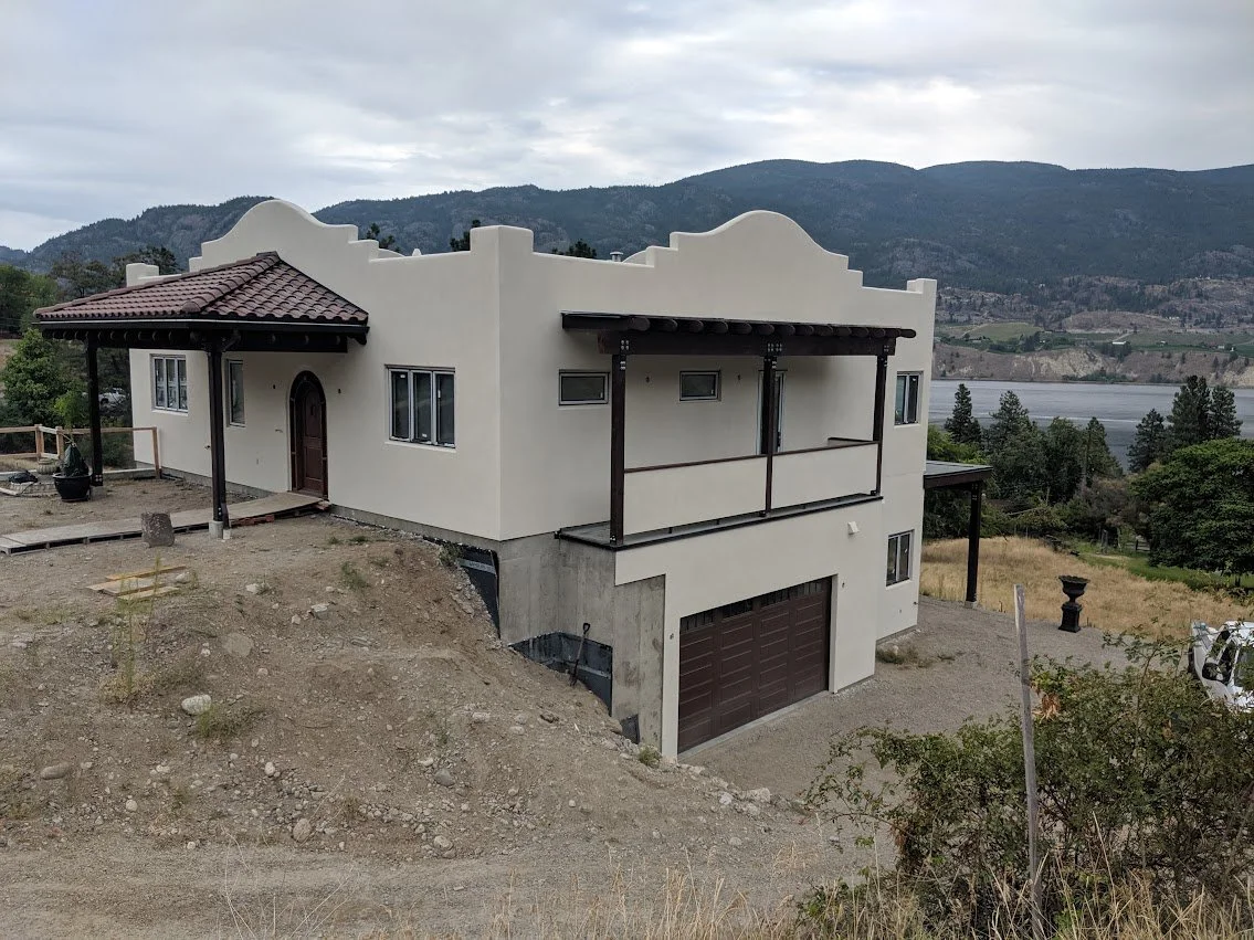 A two-story house on a sloped property with a garage, balcony, and a cream-colored exterior, surrounded by mountains and trees.