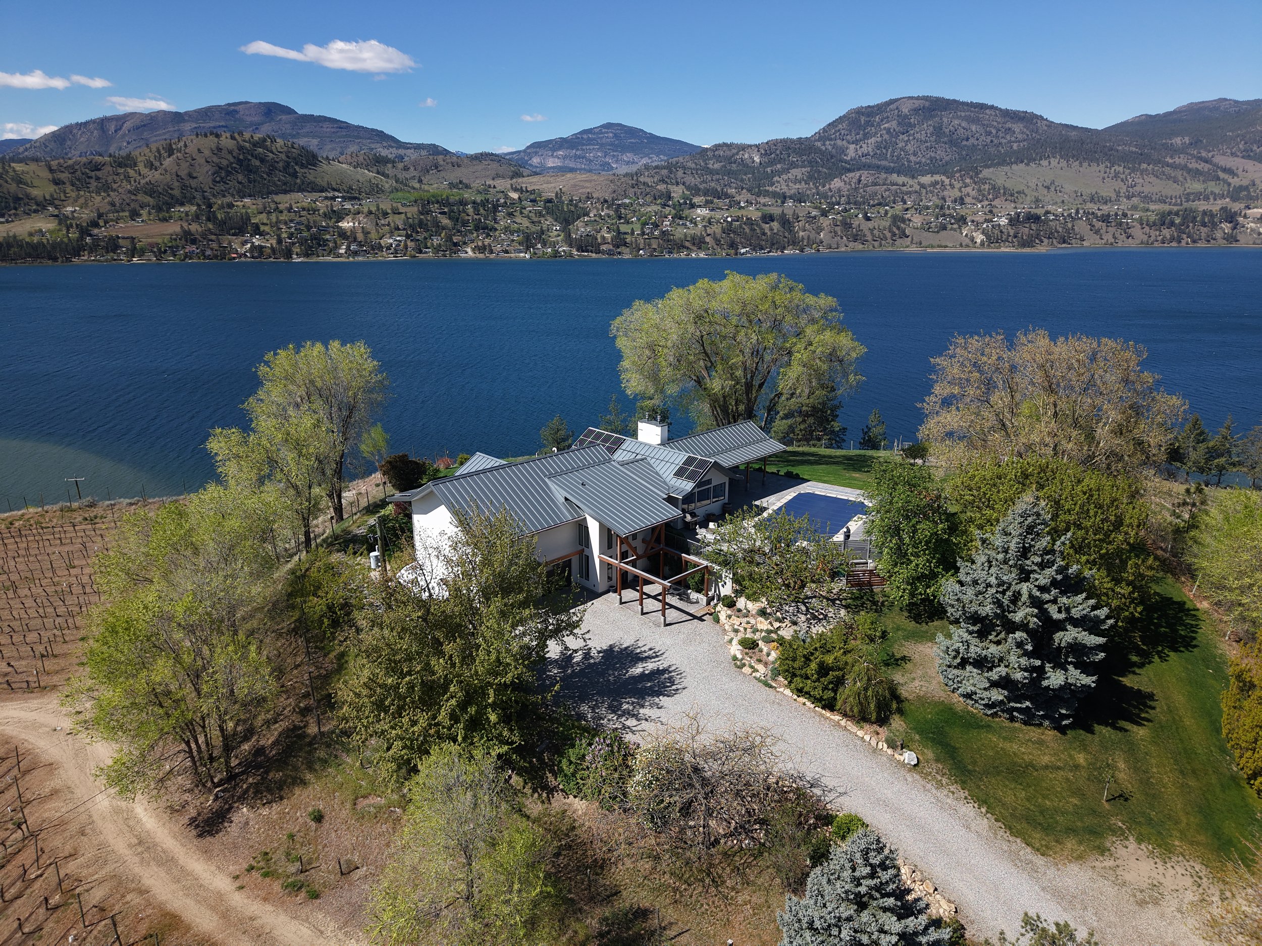 Aerial view of a lakeside home with solar panels, surrounded by trees, with a mountain landscape in the background under a partly cloudy blue sky.