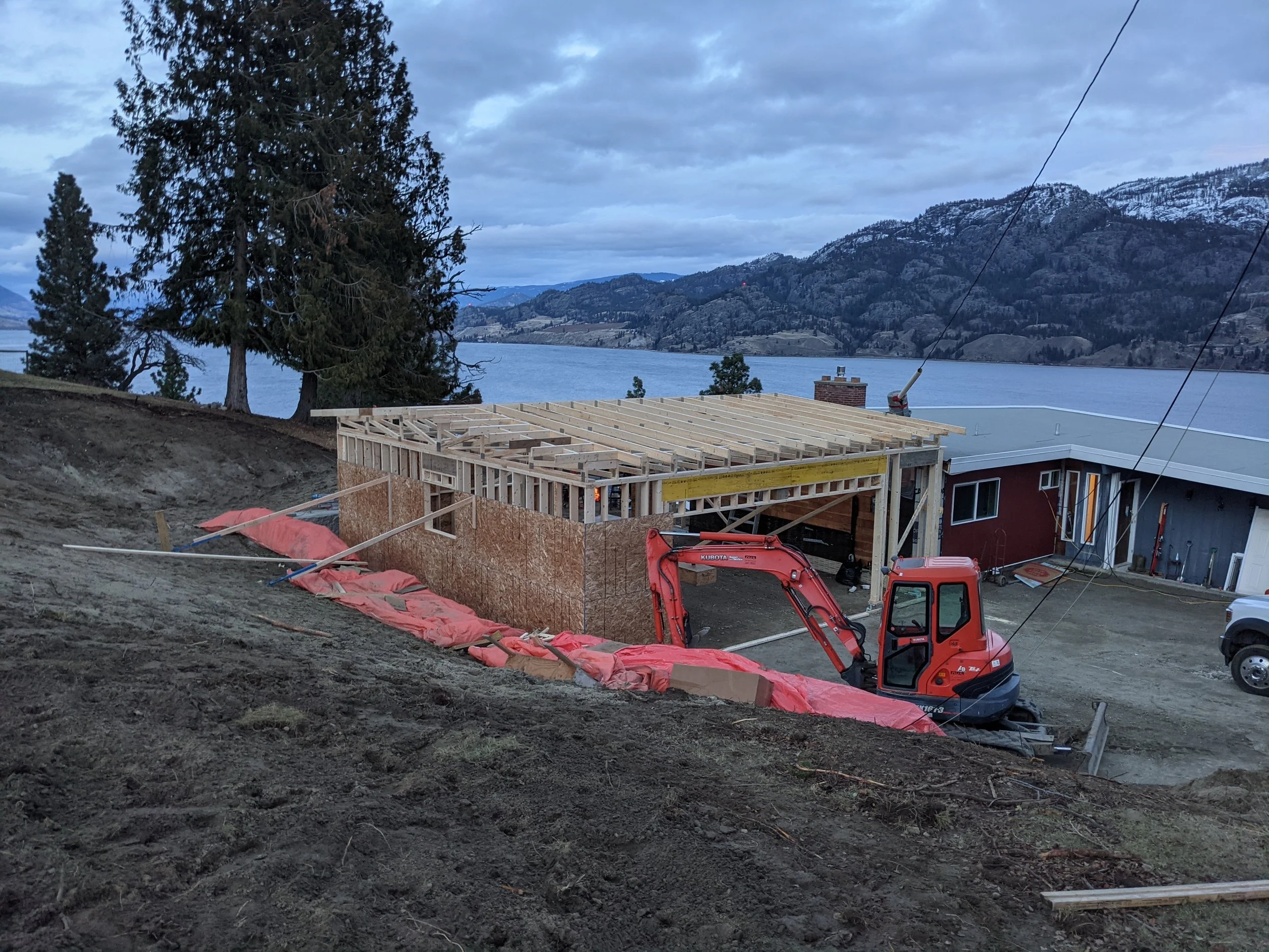 Construction site with a partially built house, a small red excavator, and scenic mountains and water in the background.