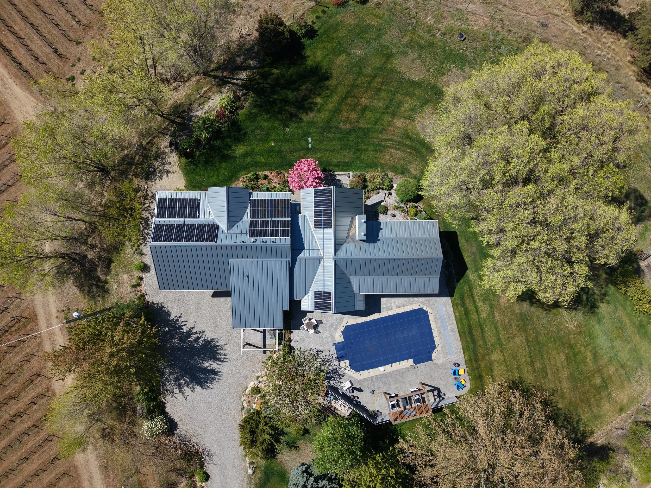 Aerial view of a house with solar panels on its roof, a backyard with trees, a lawn, and a patio area with a table and chairs, surrounded by greenery and neighboring fields.
