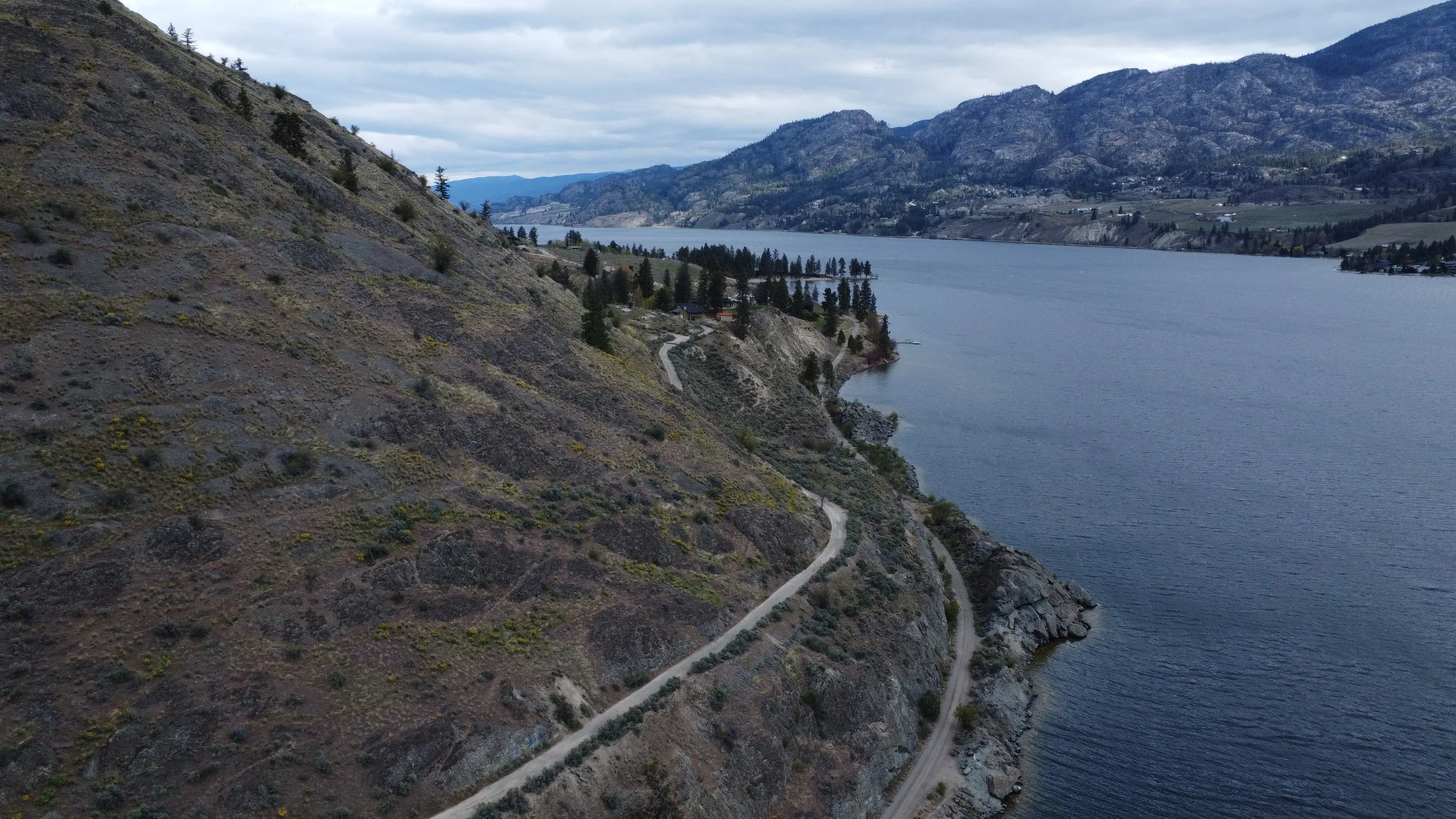 A winding dirt road along a hillside overlooking a large body of water with mountainous terrain in the background under a cloudy sky.