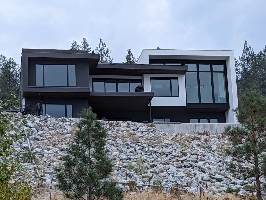 Modern multi-story house with large glass windows on a rocky hillside, surrounded by trees.
