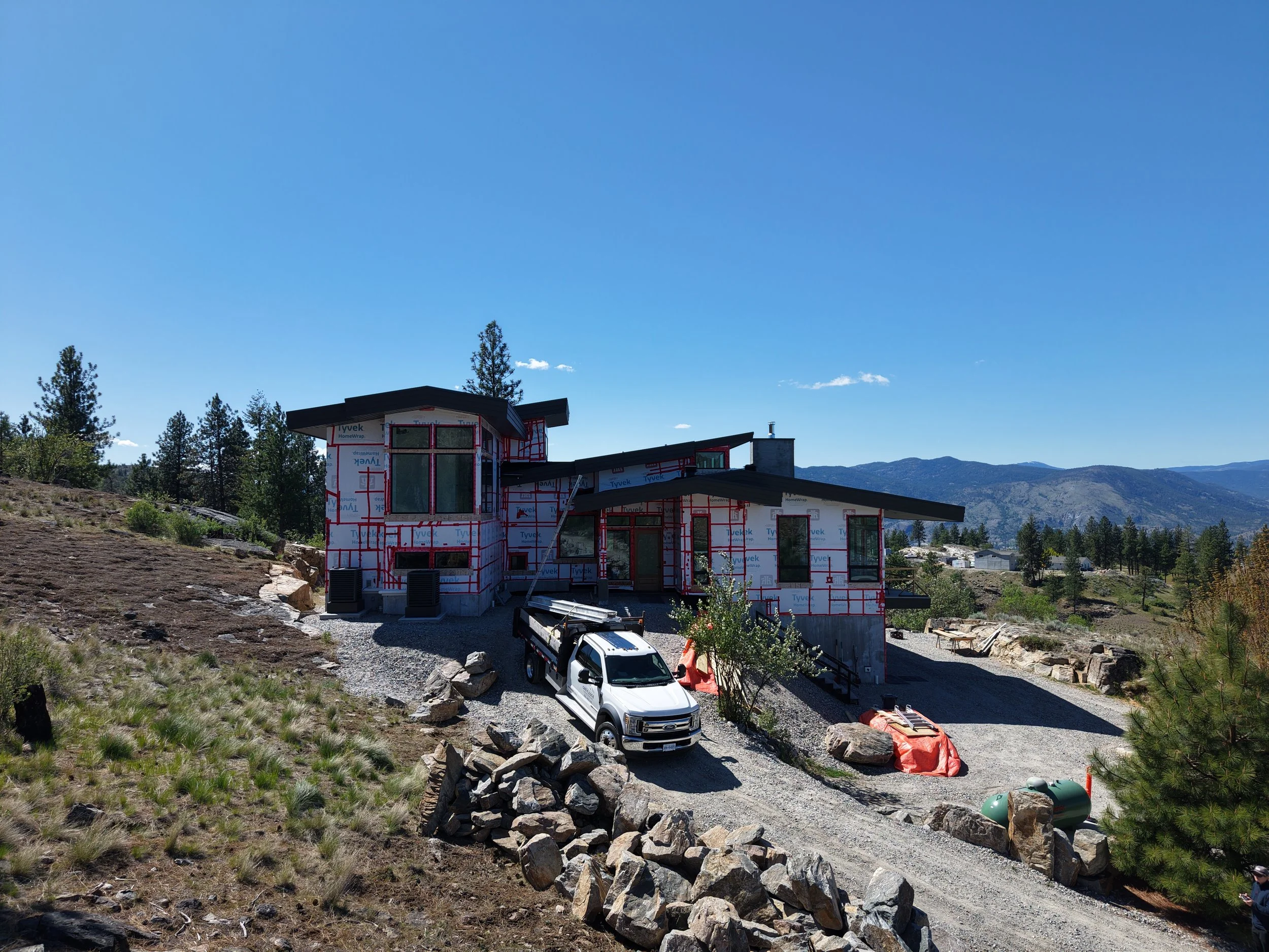 A modern house under construction in a mountainous area, with a black roof, large windows, and wrapped in Tyvek building wrap. A white pickup truck and construction materials are in the foreground, with trees and mountains in the background.